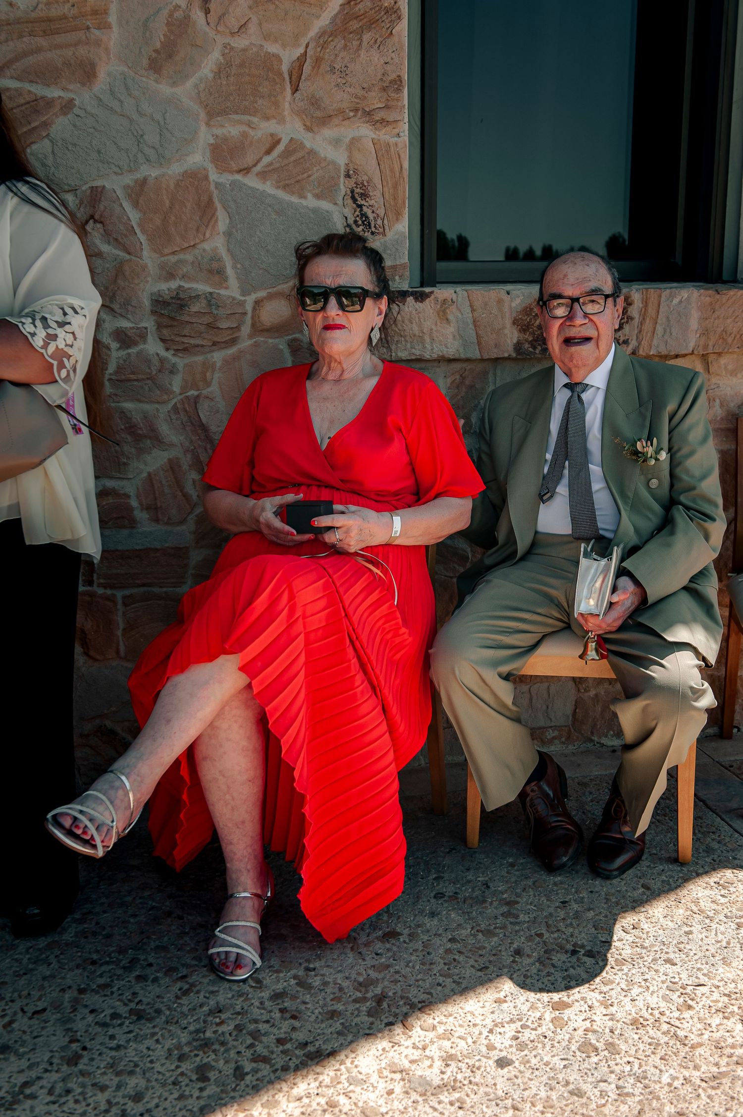Two people sit on chairs against a stone wall, one wearing a bright red dress and the other in formal attire.