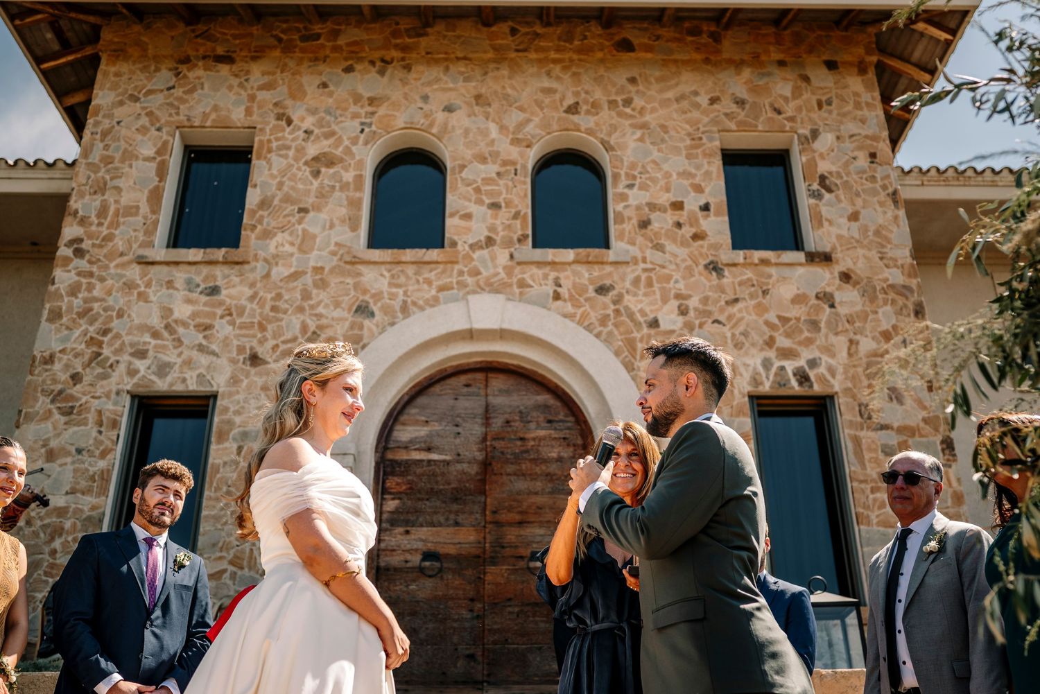 Wedding guests mingle and socialize in front of a rustic stone building during an outdoor reception.