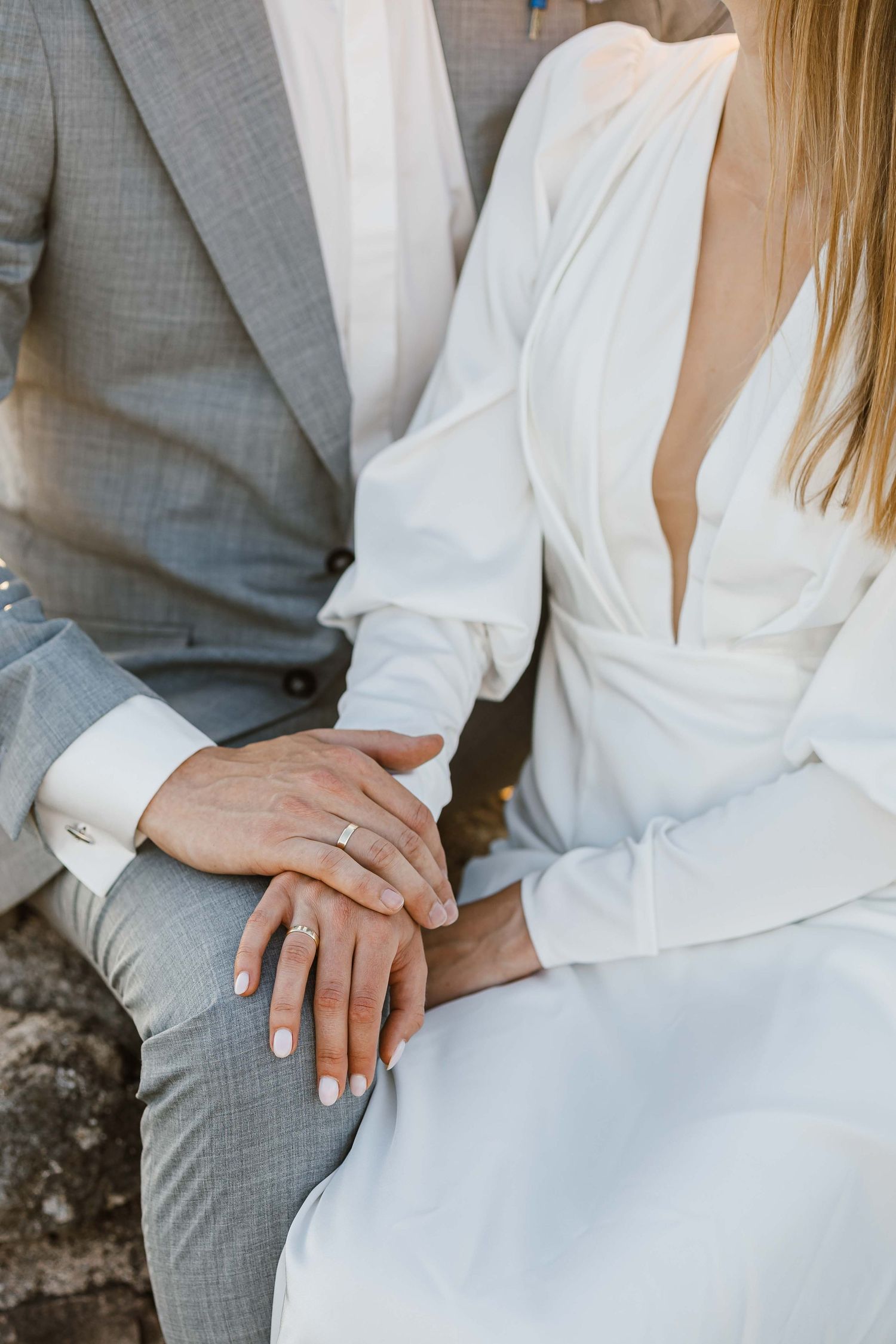 Couple's hands with wedding rings against grey suit and white dress backdrop.