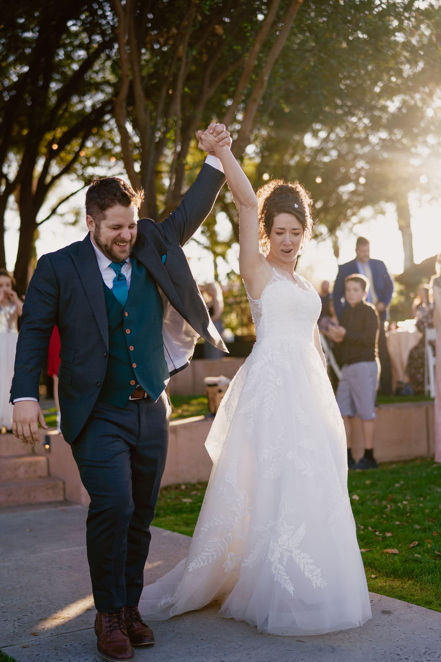 Newlyweds celebrate with raised hands while walking together outdoors in evening light.