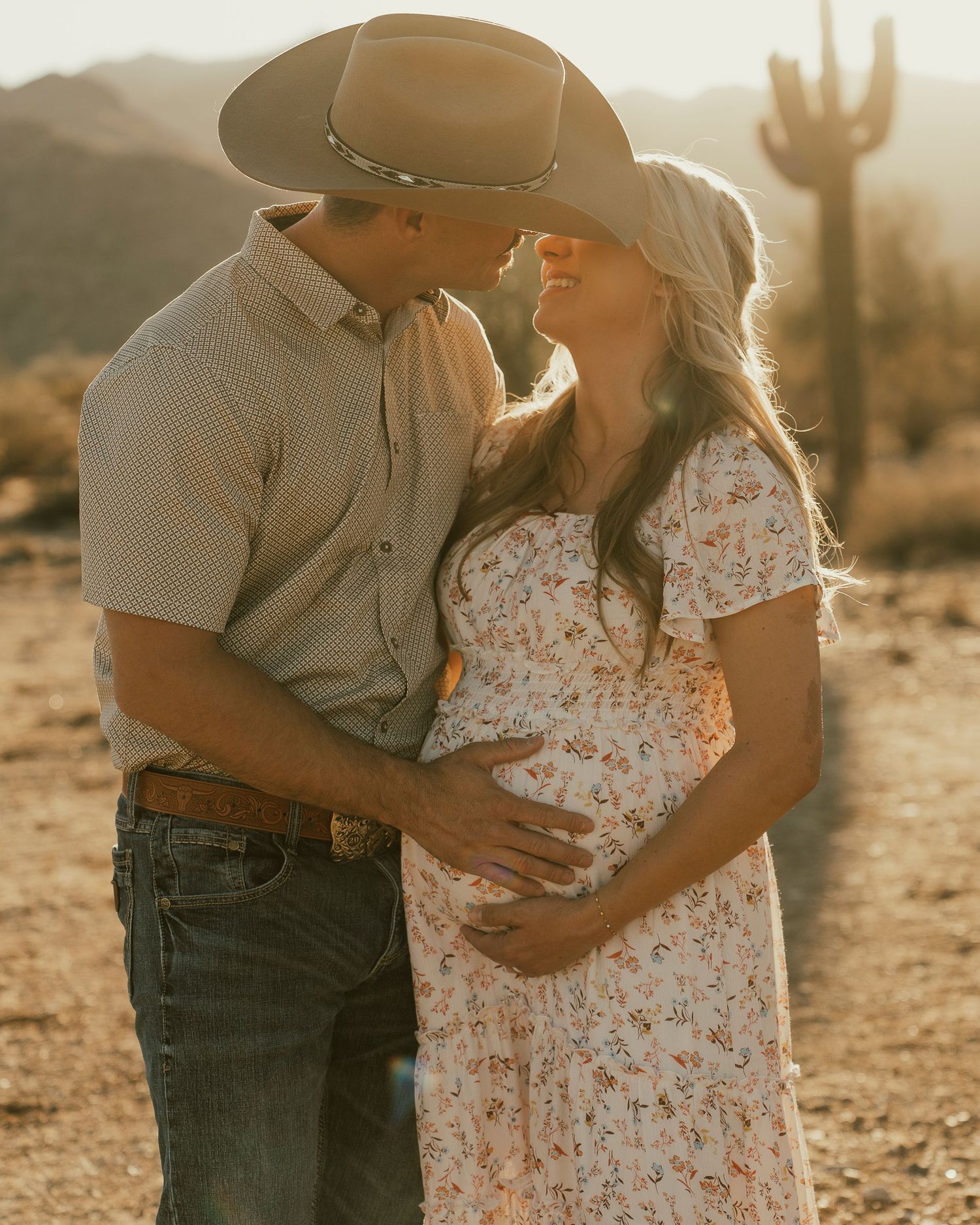 Intimate desert maternity photo with couple in western wear against saguaro cactus backdrop.