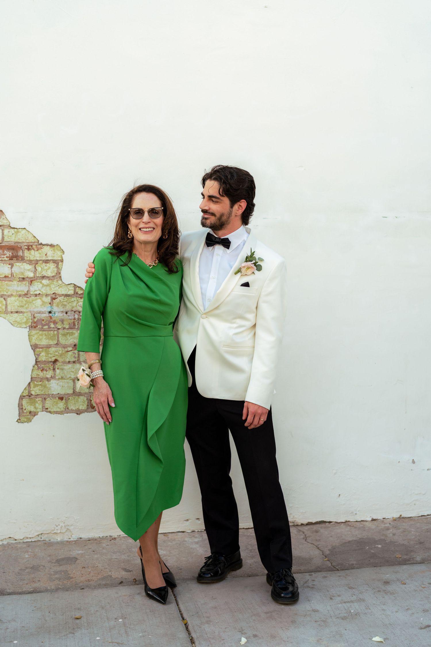 Person in emerald green dress poses alongside companion in white jacket against white wall.