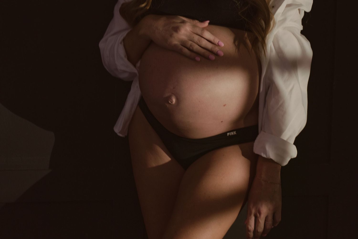 Woman in white shirt and black lingerie poses seductively against dark background in boudoir photoshoot.