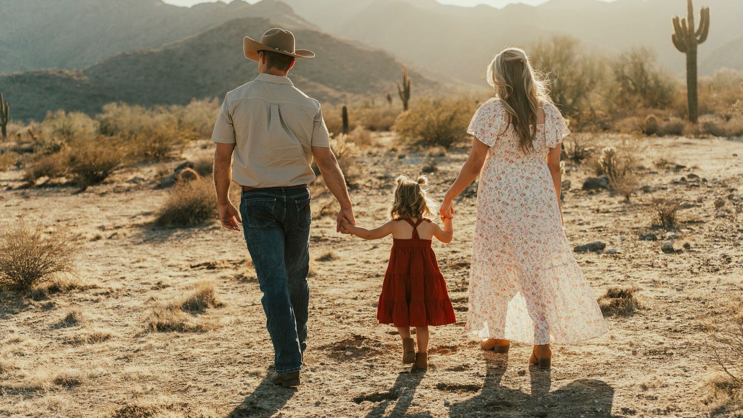 A family walks hand in hand through a desert landscape with mountains and cacti.