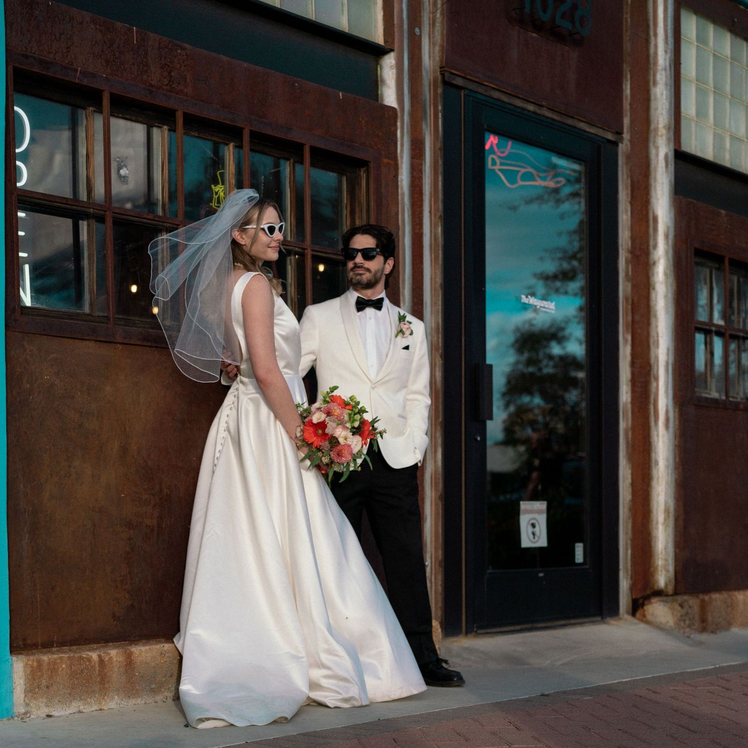 Wedding couple in formal attire pose outside a brick building with the bride holding a vibrant bouquet.
