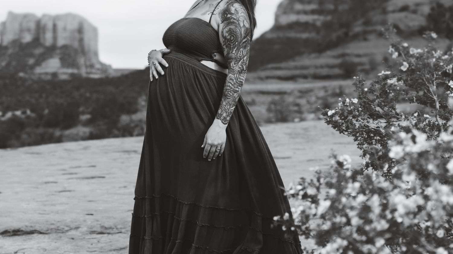 Black and white photo of a pregnant woman in a long dress standing on rocky terrain with desert mountains in background.