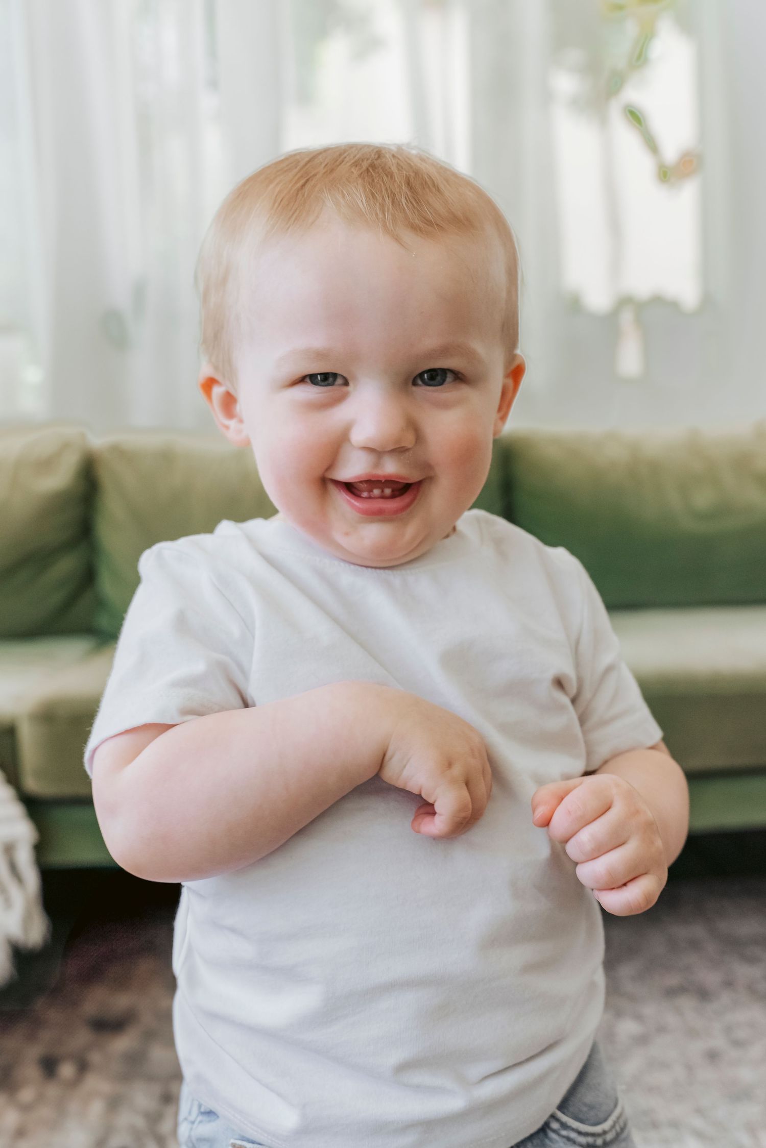 Smiling toddler in white t-shirt standing against green couch in bright room.