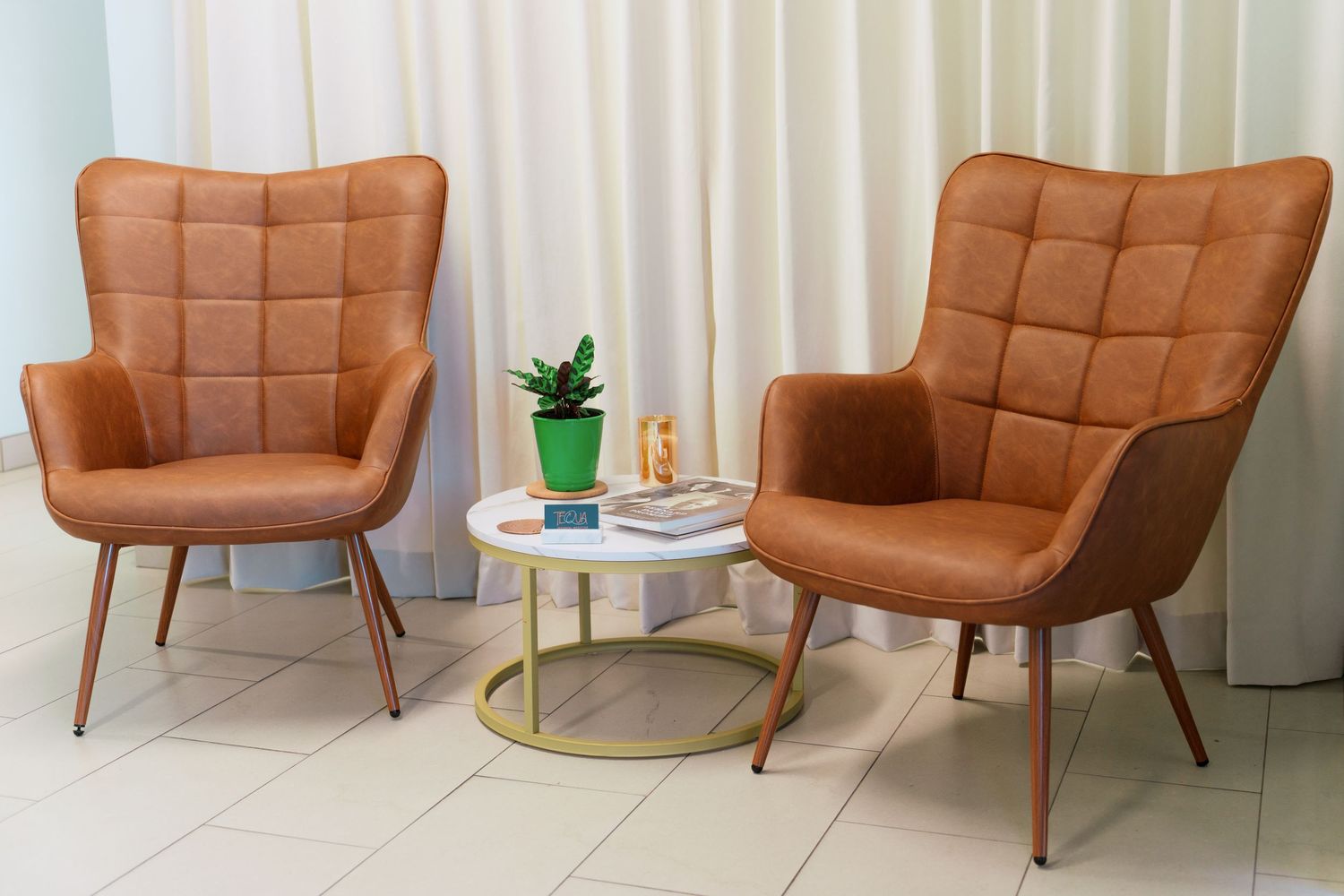 Two brown tufted accent chairs with wooden legs flank a small round side table with a plant in a modern living space.