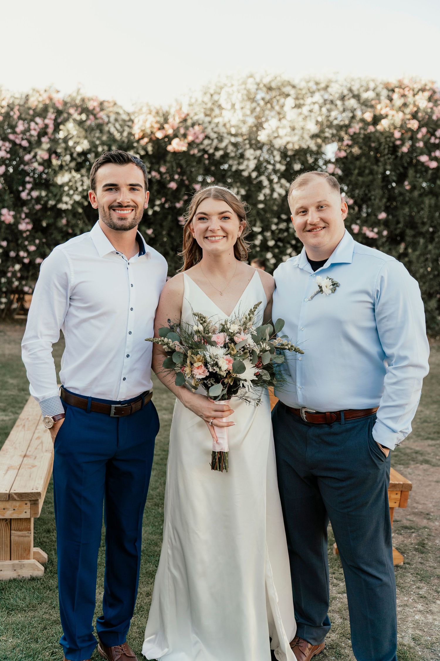 Three people pose outdoors with a floral bouquet against a blooming garden backdrop.