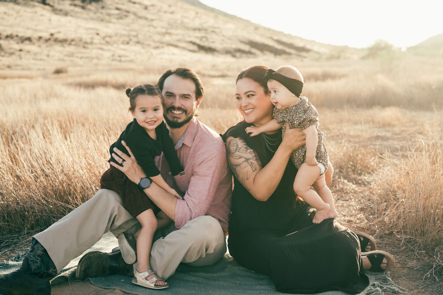 A family sits together in a golden field during sunset with mountains in the background.