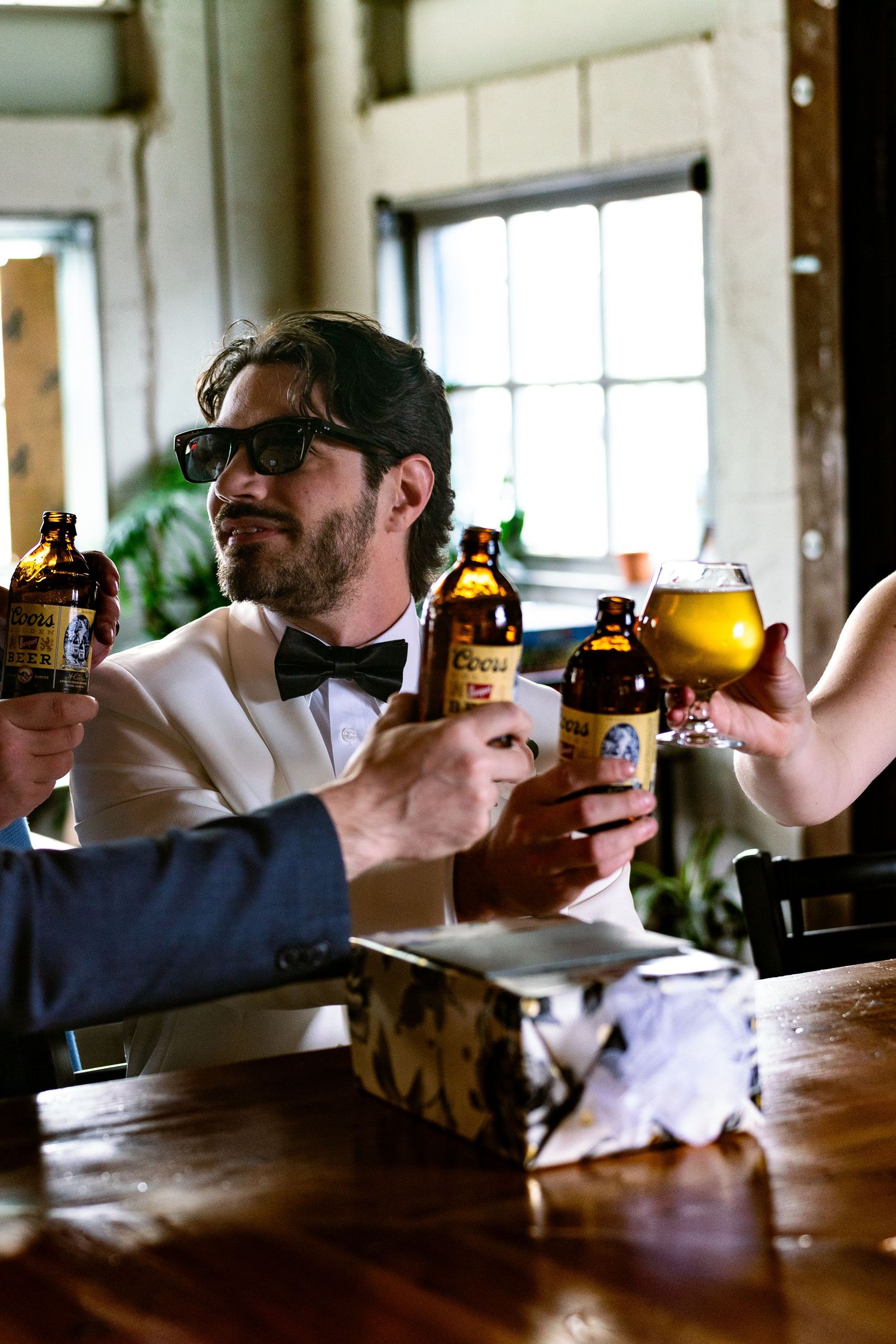 Wedding party toasting with beer bottles in rustic indoor setting.