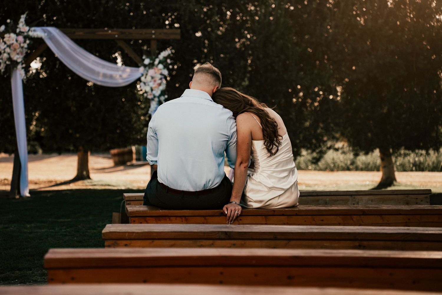 Couple sits together on wooden benches under a decorated wedding arch at sunset.