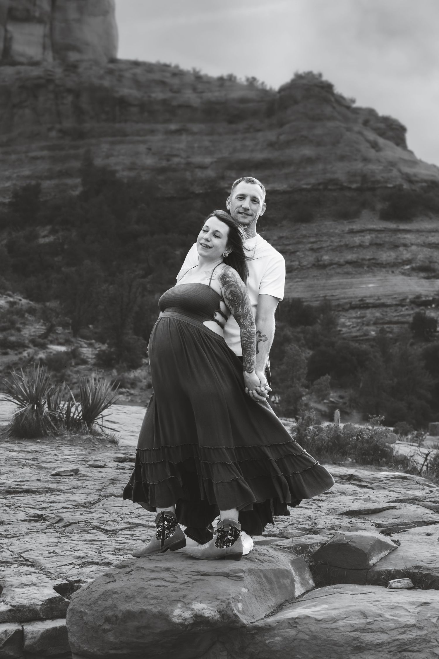 A romantic black and white photograph of a couple dancing together on rocky terrain with desert cliffs in the background.