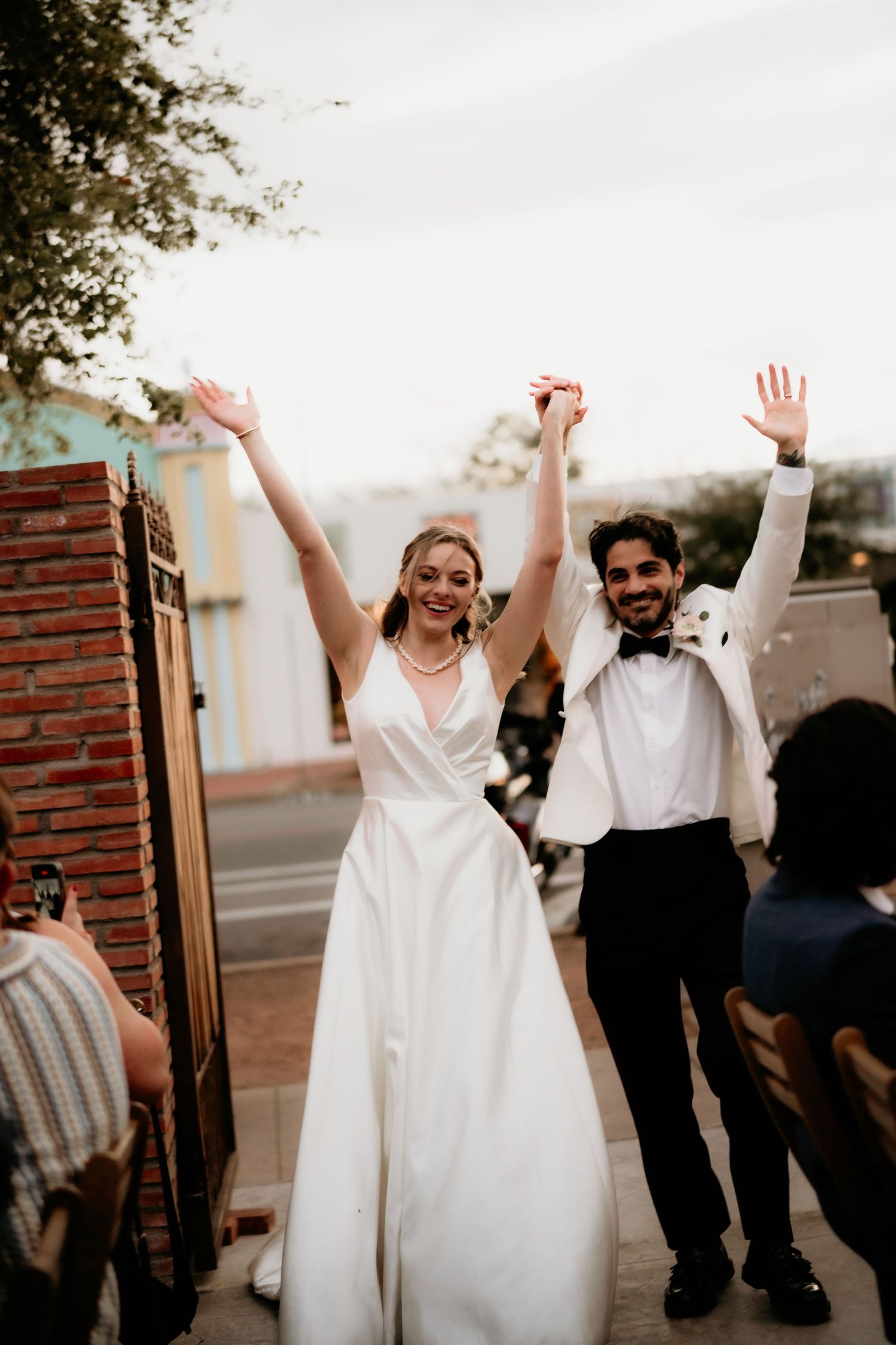 A joyful wedding couple raises their arms in celebration during their outdoor reception.