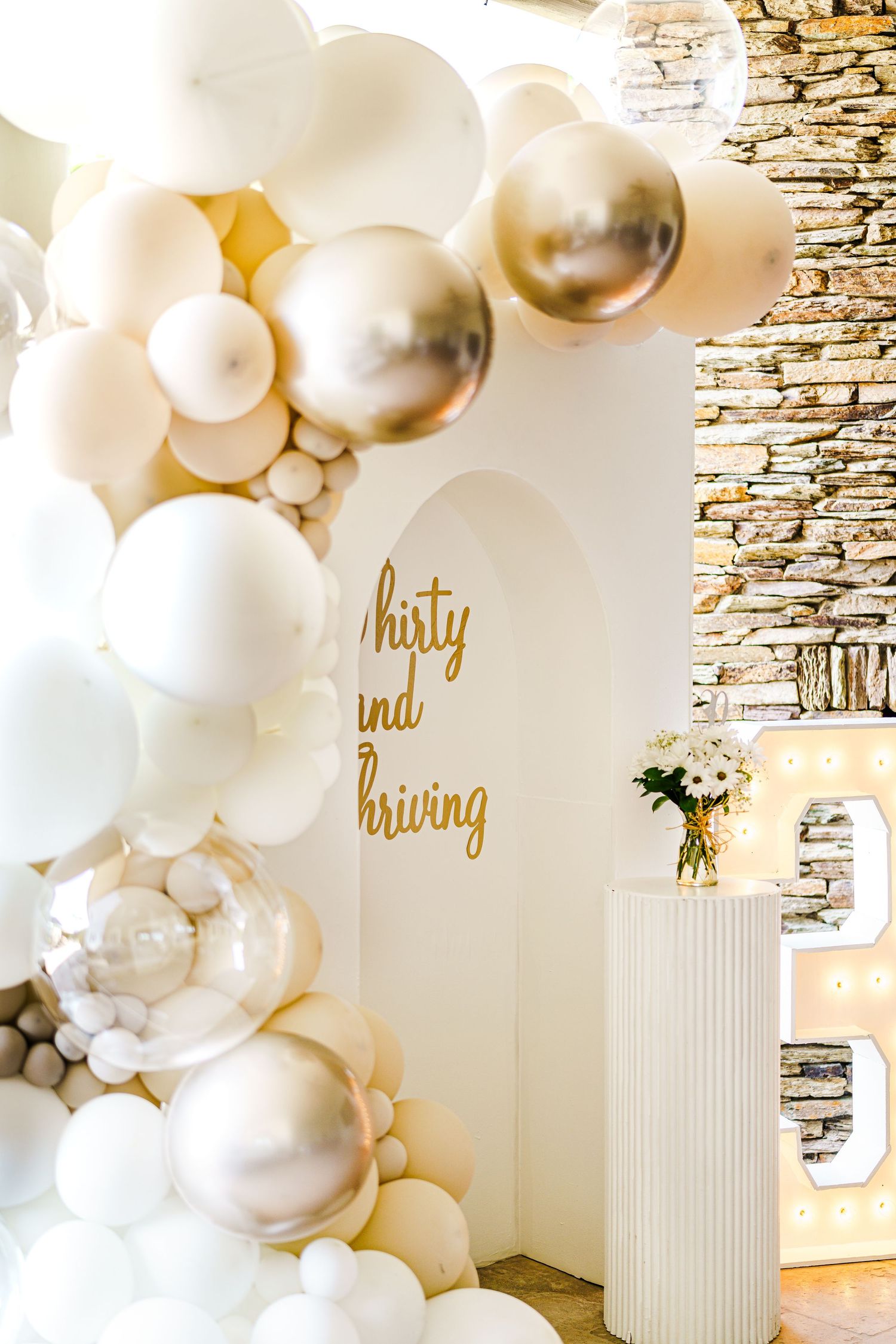 Elegant party backdrop with white and gold balloon arch against stone wall. Session in Phoenix Arizona