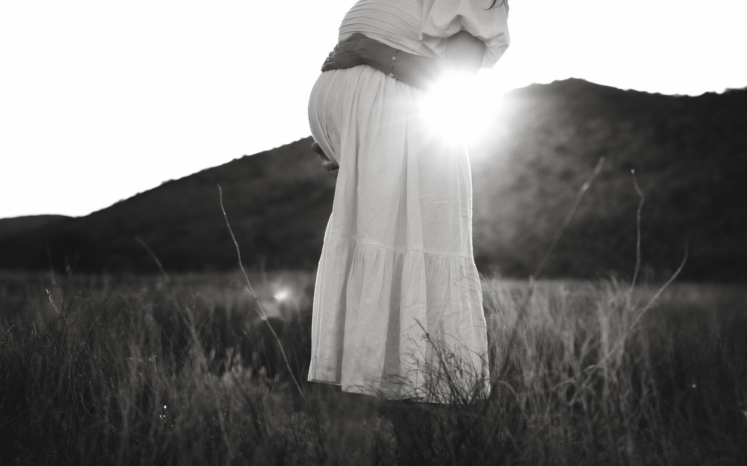 Black and white silhouette of pregnant woman in flowing dress against sunlit landscape.