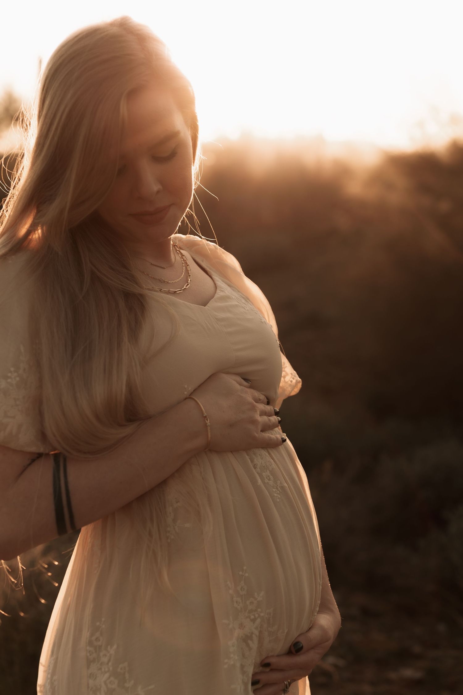 Expectant mother in white dress caresses her baby bump during golden hour sunset maternity photoshoot.