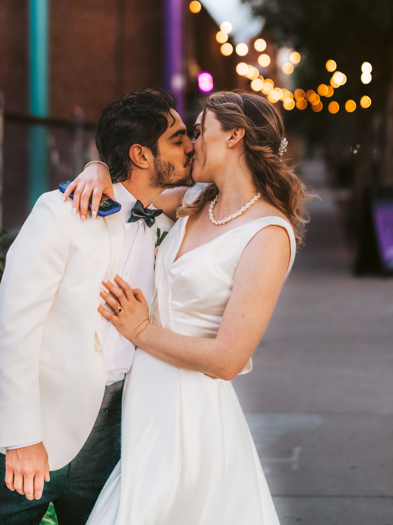 Romantic wedding couple sharing a kiss in white formal attire under evening string lights.