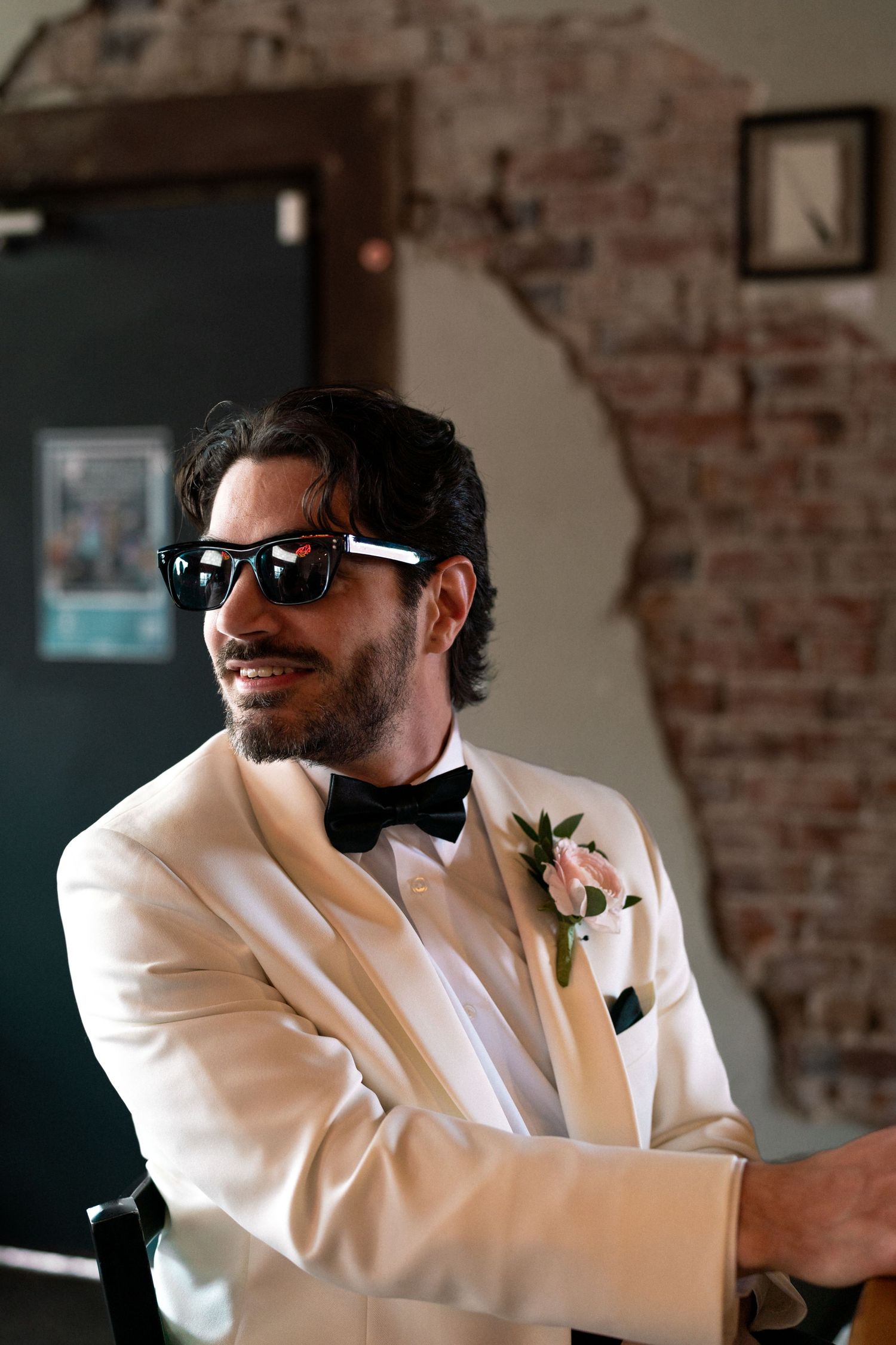 Groom in white tuxedo jacket and sunglasses smiling confidently against brick wall.