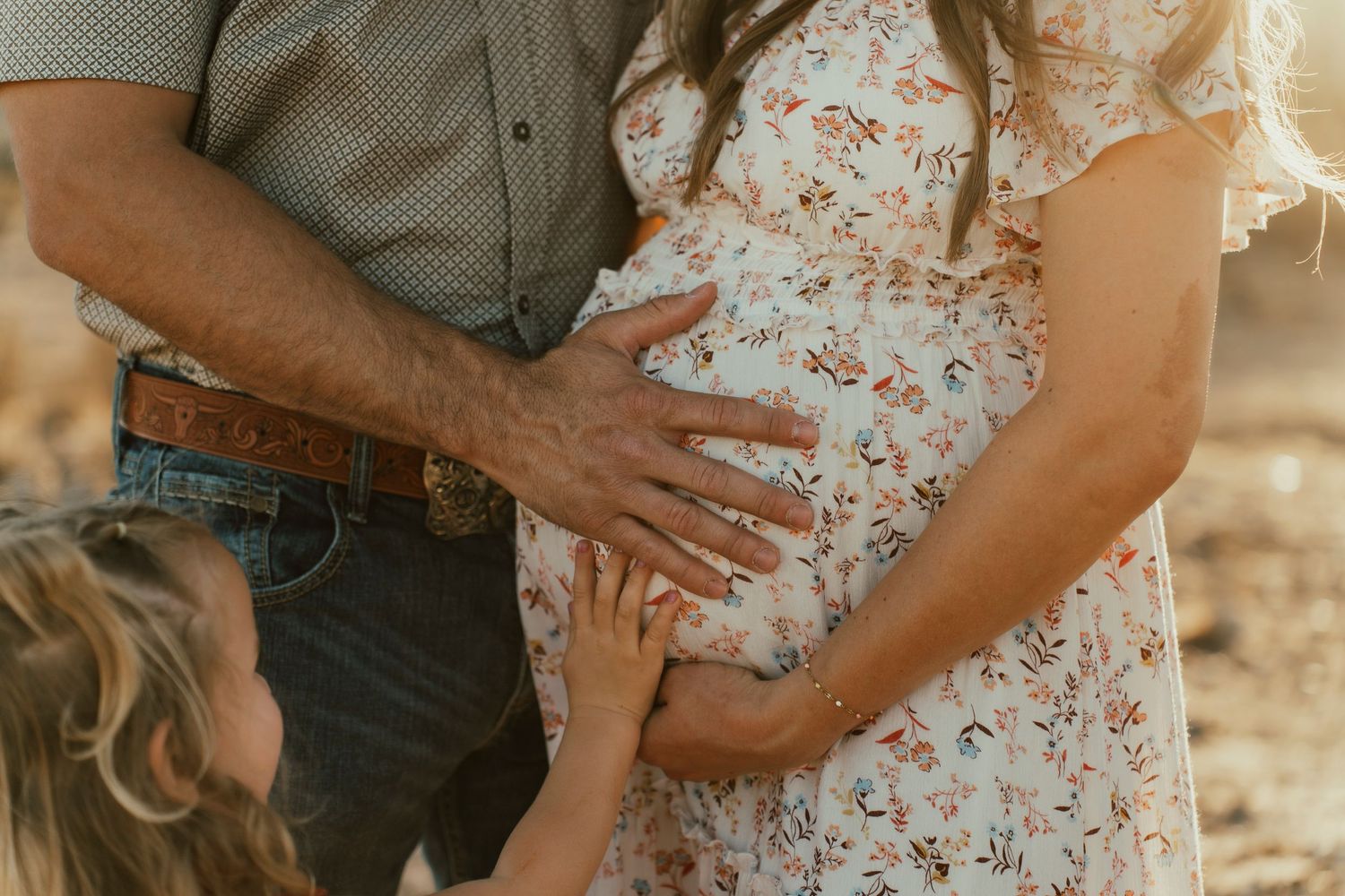 Close-up of family hands resting on pregnant belly in desert setting.