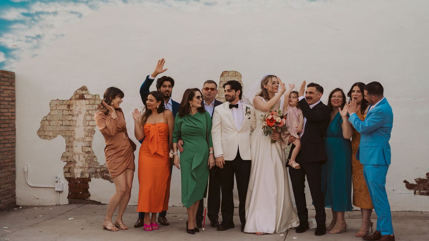 Wide panoramic shot of wedding party and guests gathered against brick wall outdoors.