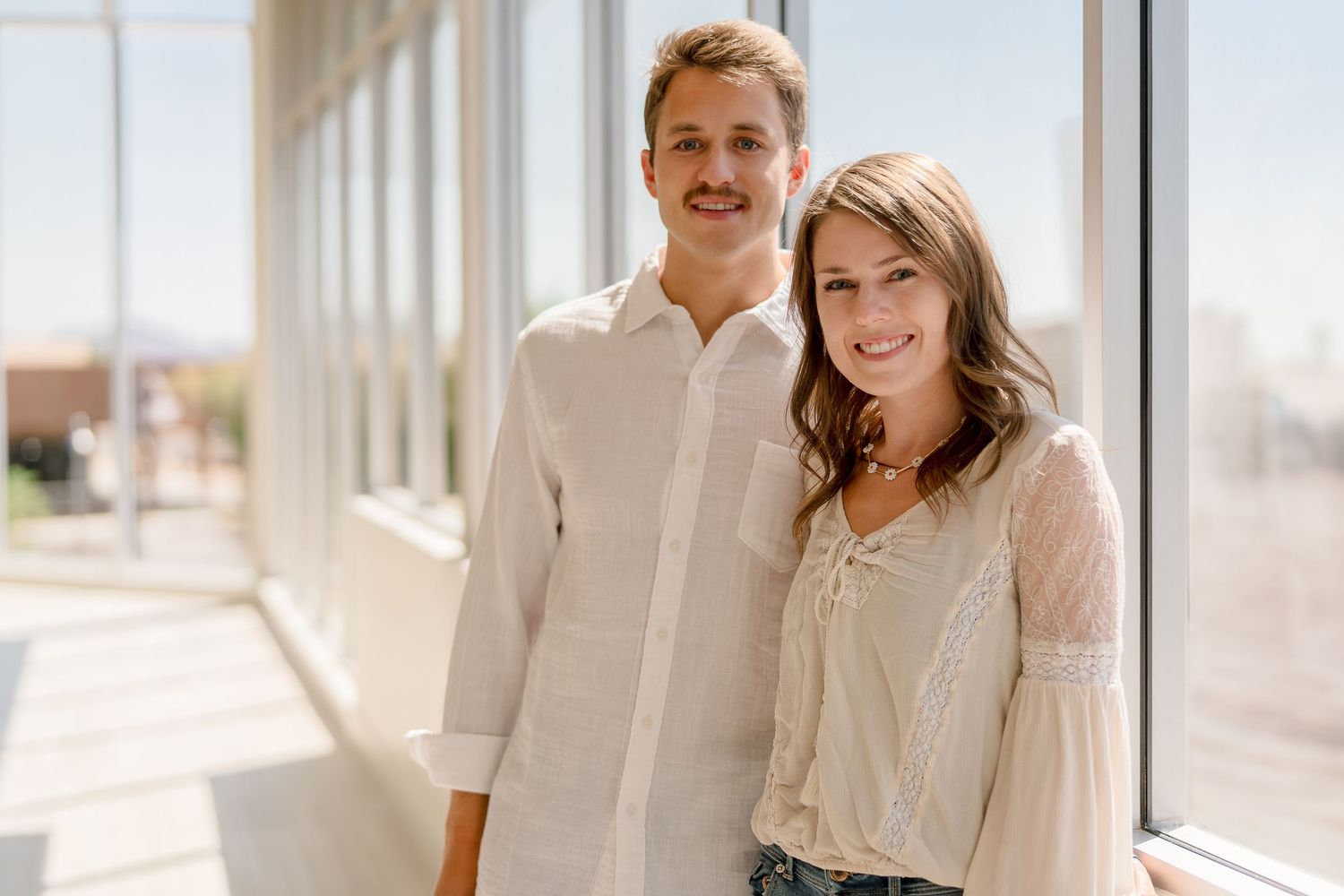 Two people stand together in casual attire against a bright white exterior wall.