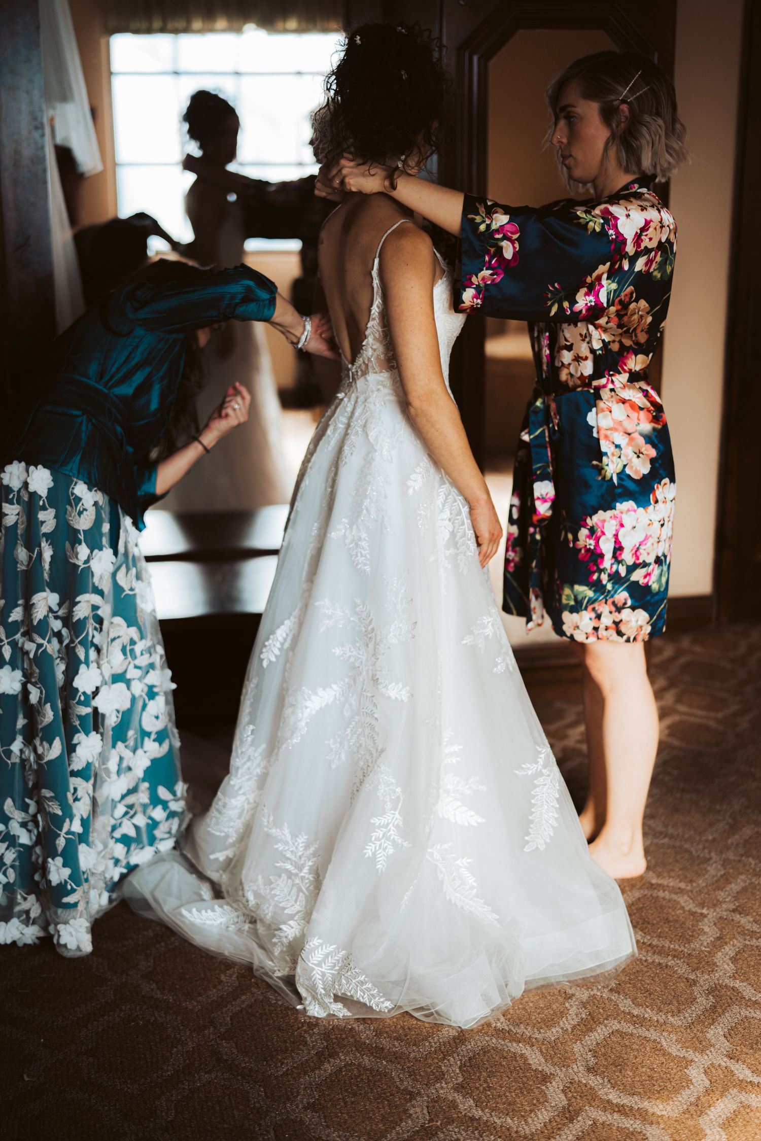 Women helping with final wedding dress preparations in a dimly lit room with floral dresses and white gown.