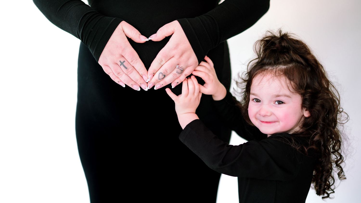 A young child with dark curly hair wearing black clothing holds up a pink ribbon while posing against a white backdrop.