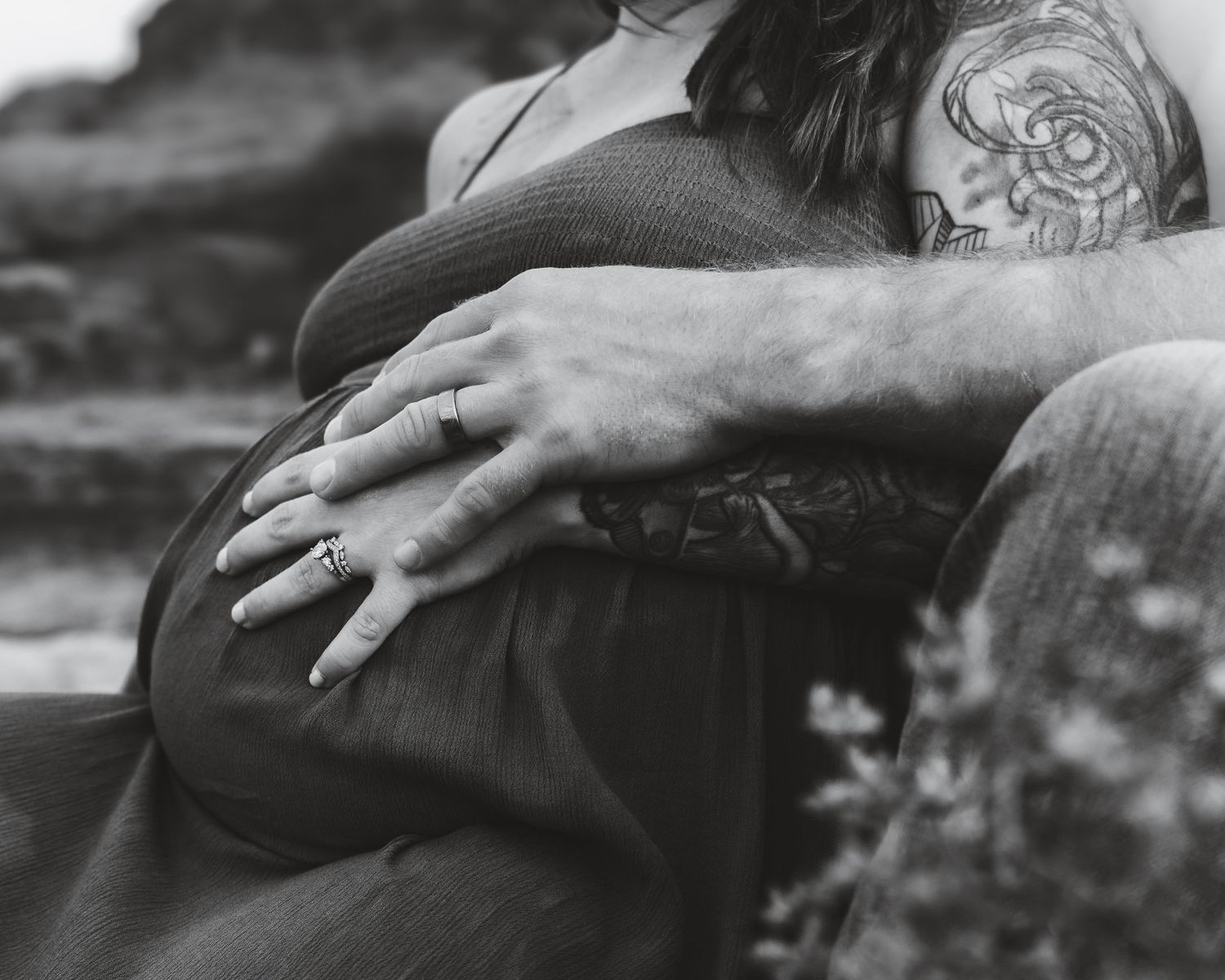 Black and white close-up photo of hands resting on a pregnant belly with visible tattoo artwork.