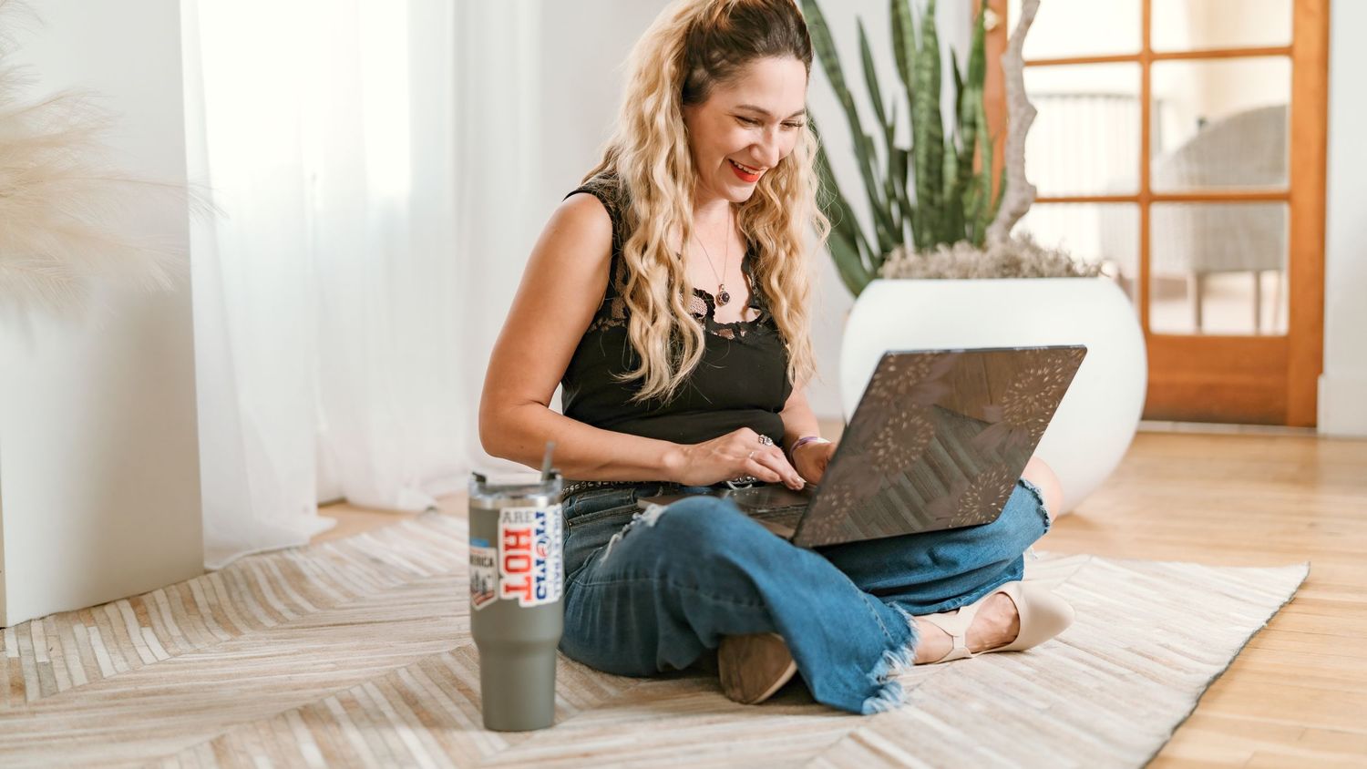 Woman sitting on white bed while using laptop and tablet devices in bright modern bedroom.