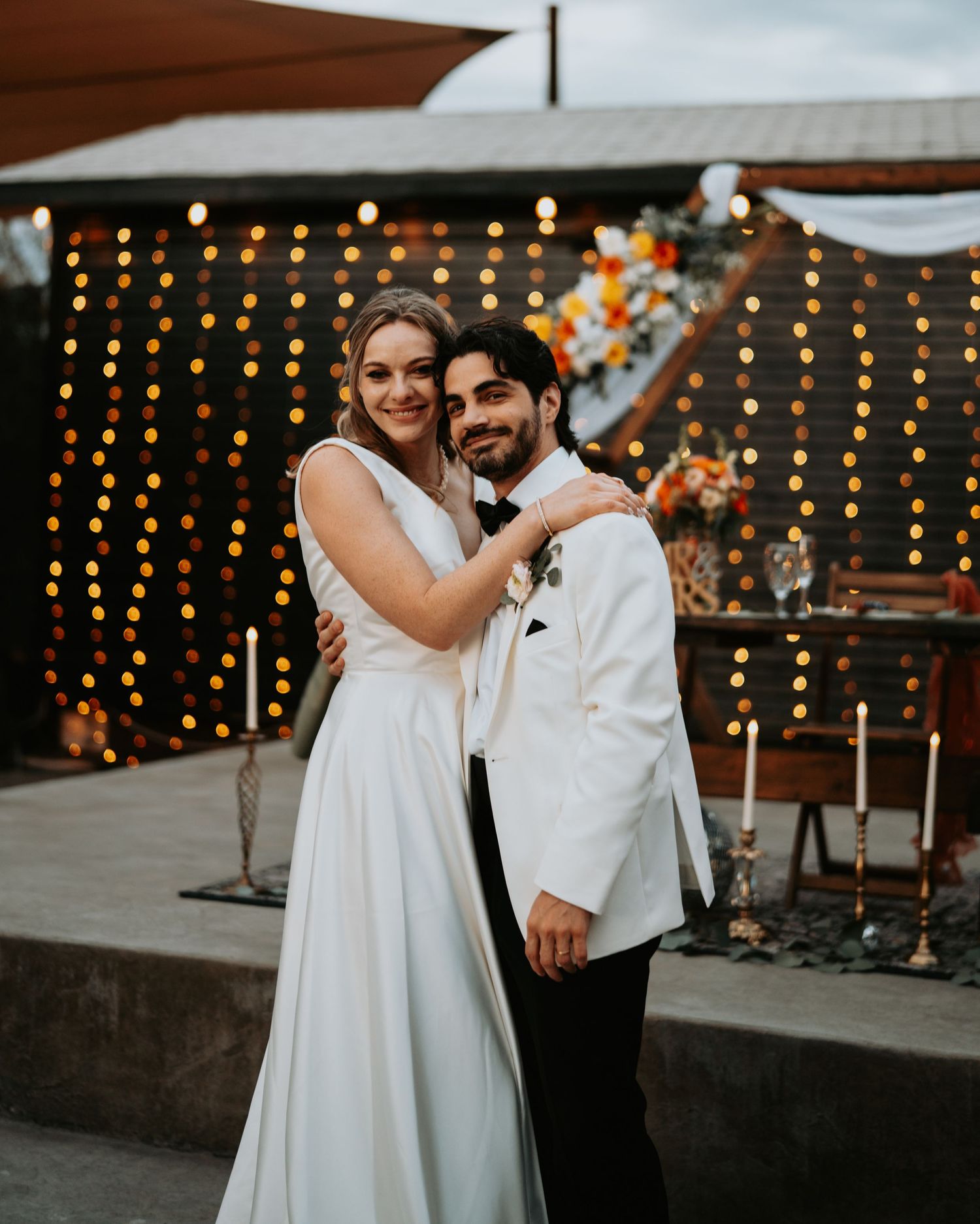 Romantic couple embracing under string lights at an outdoor wedding reception.