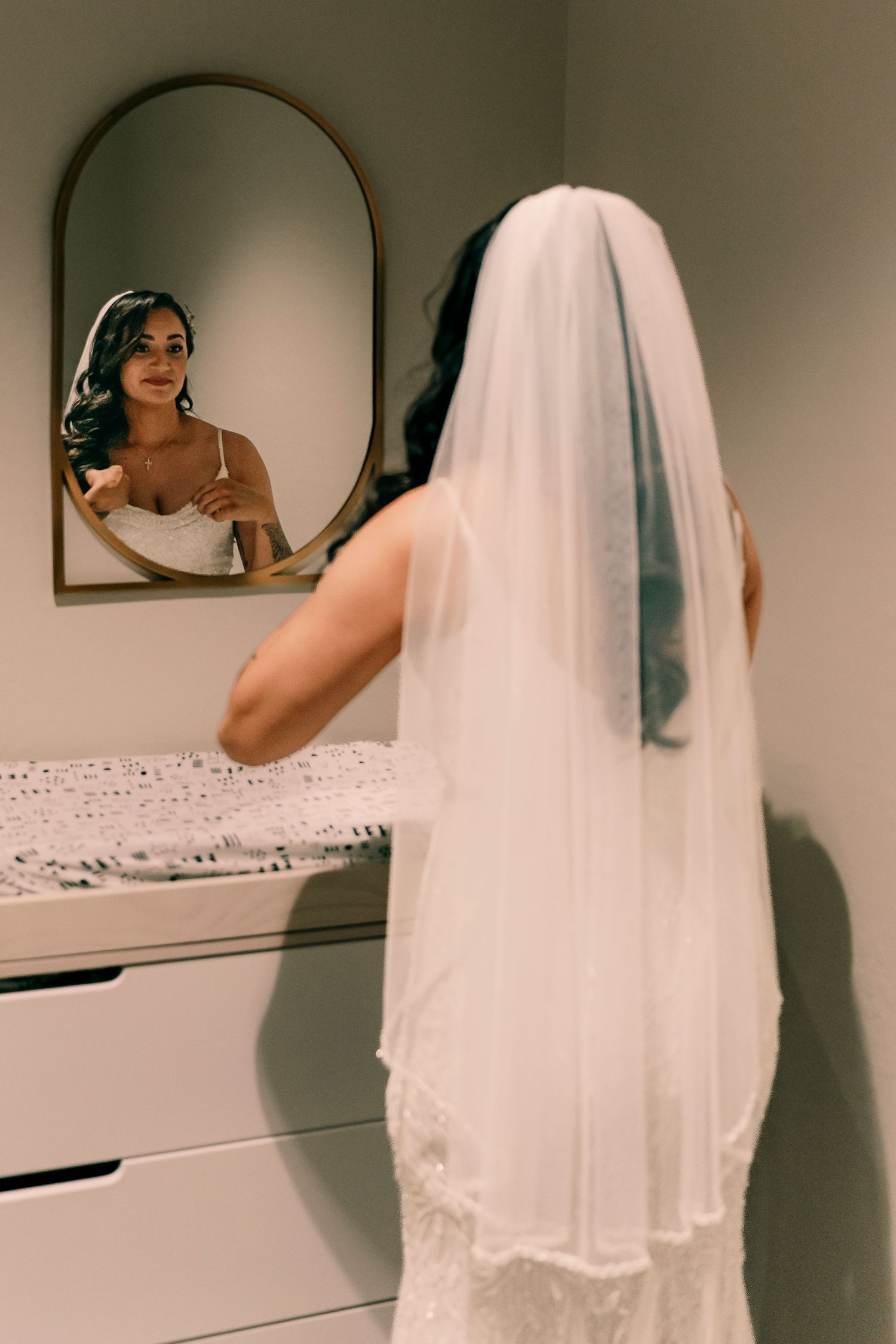 A bride admires her reflection in an arched mirror while wearing a flowing veil.