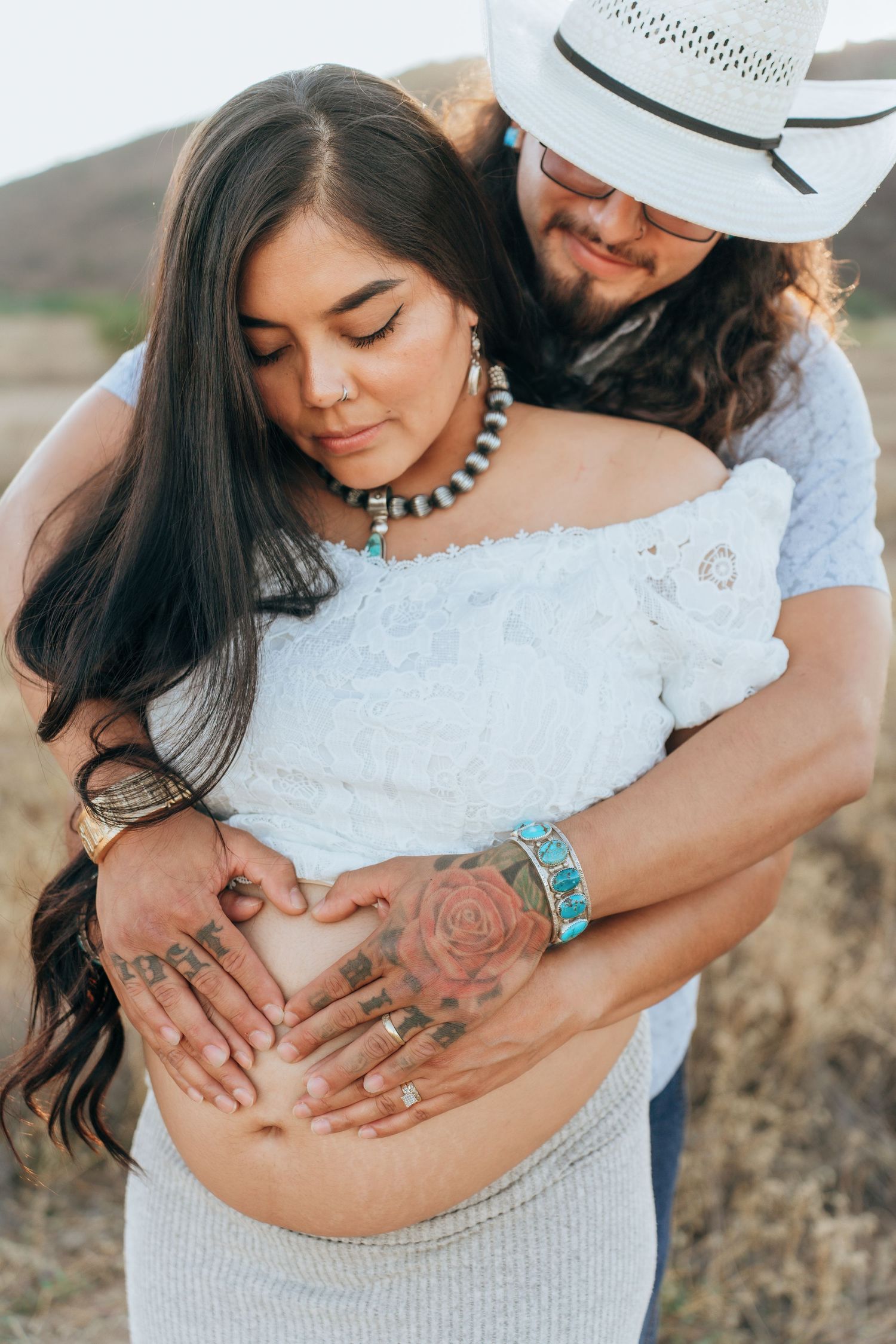 Close-up shots of hands with tattoos resting on a pregnant belly, wearing turquoise jewelry and white lace dress.