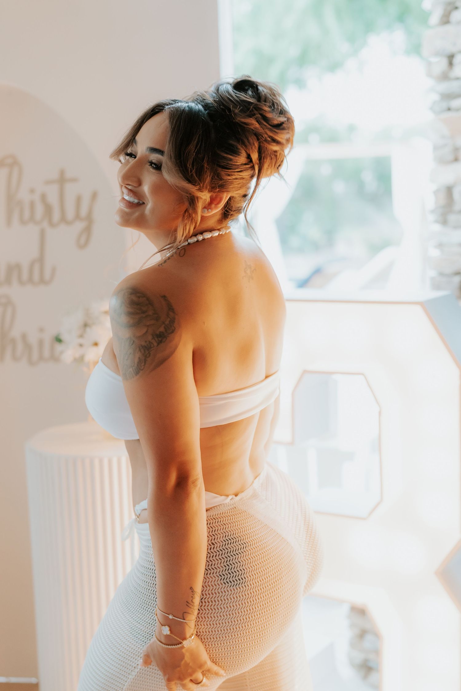 Woman in white swimwear poses confidently against a bright interior backdrop with visible tattoo detail.