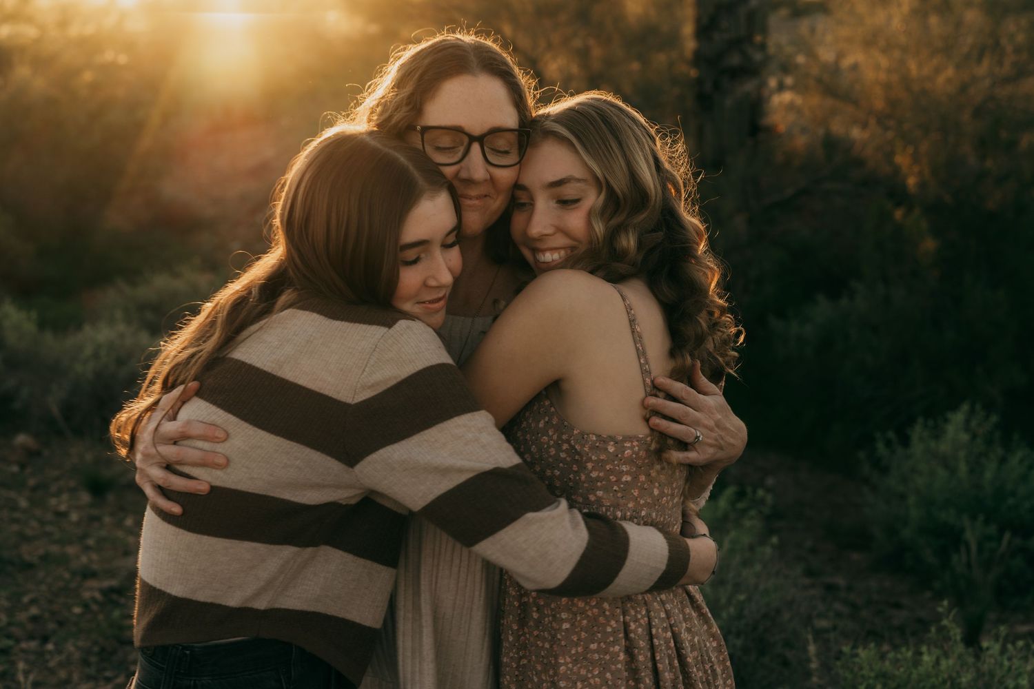 Three people share an intimate embrace outdoors during golden hour with sunlight filtering through autumn trees.
