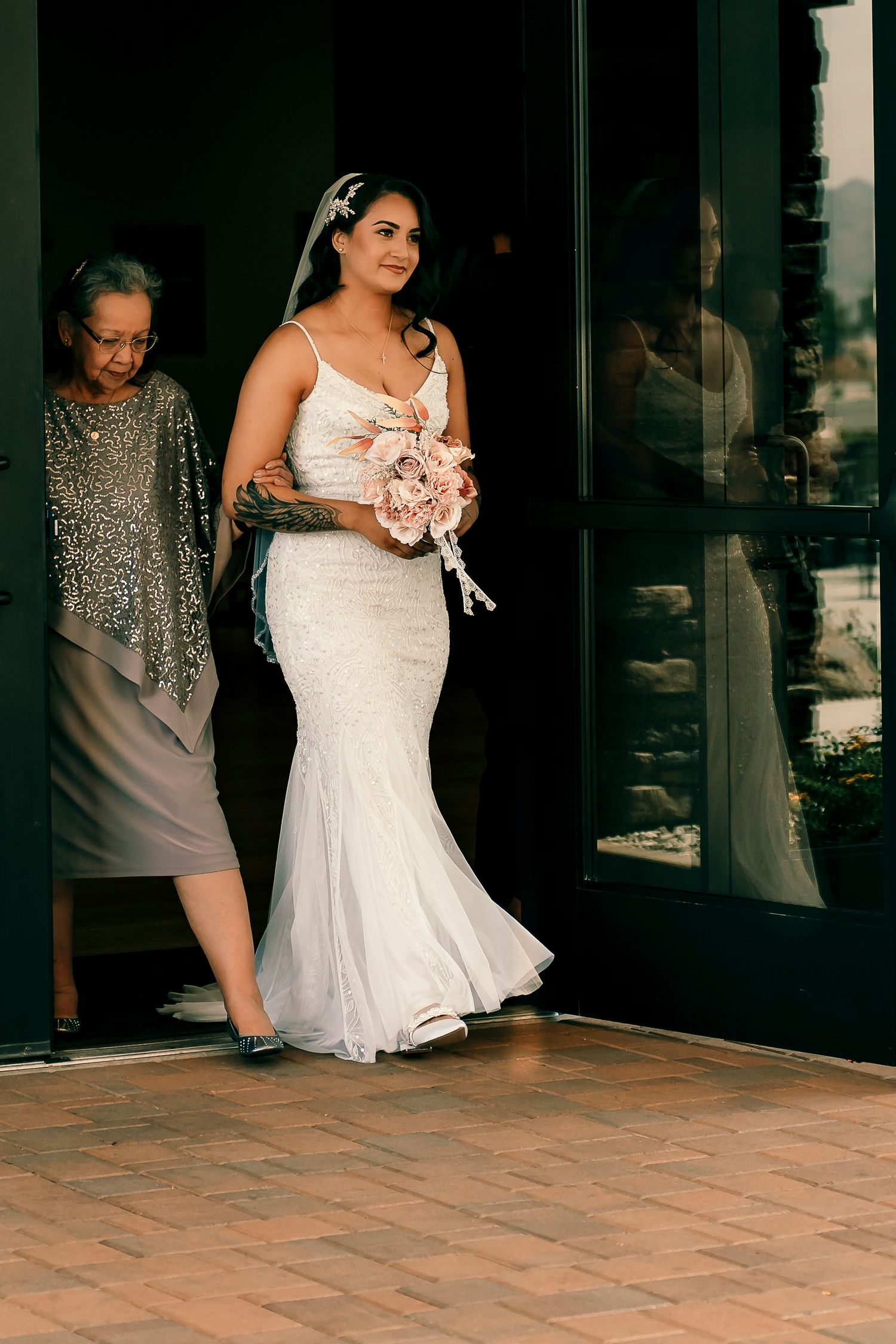 A woman in a fitted white wedding dress holds a pink bouquet while walking on a brick patio.