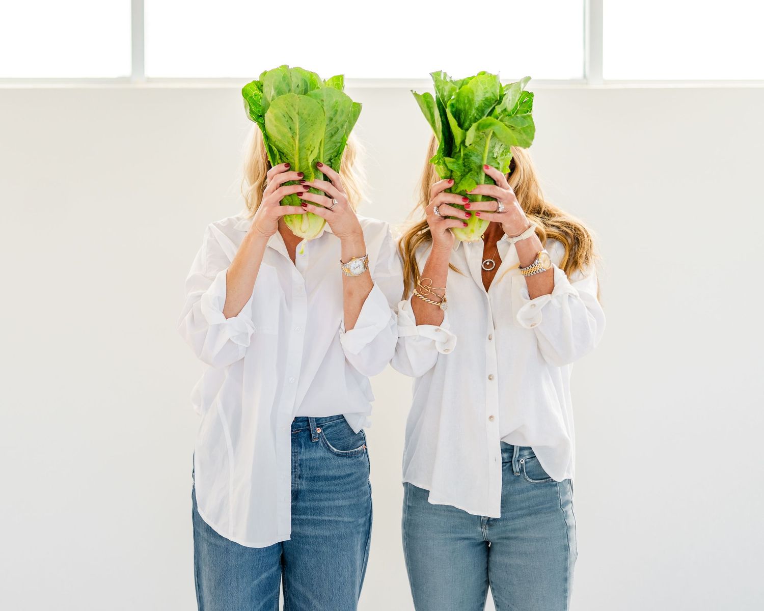 Two people in white shirts and jeans hold green lettuce heads in front of their faces against a white background.