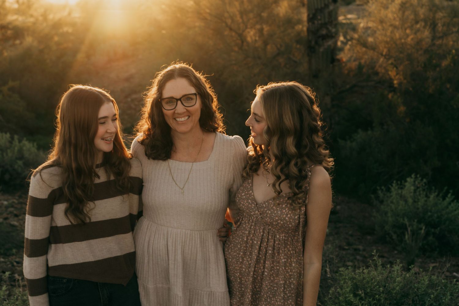 Three friends stand together smiling in warm sunset light with golden backlit glow through autumn trees.