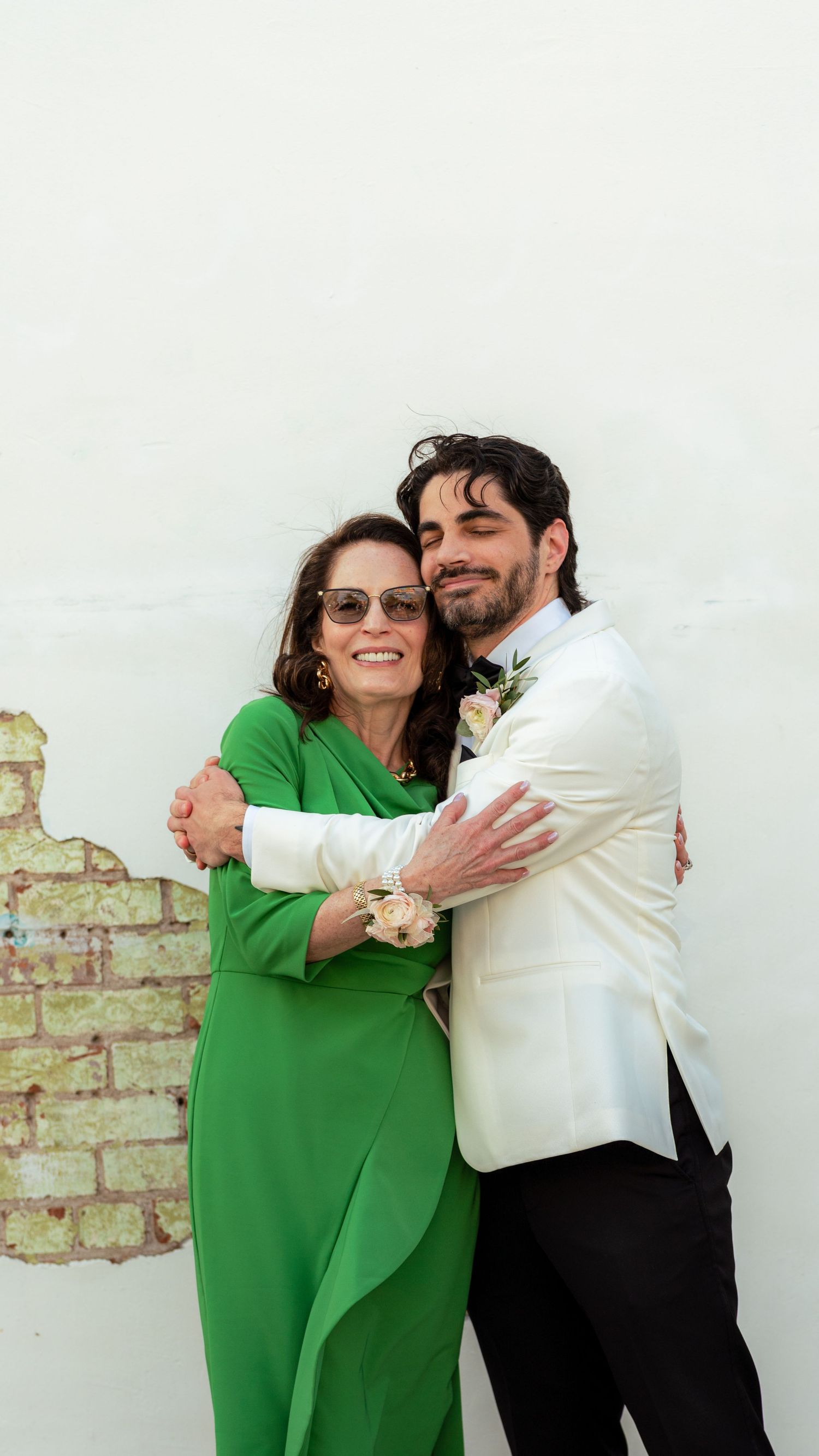 Two people embrace against white wall, one wearing vibrant green dress and sunglasses.