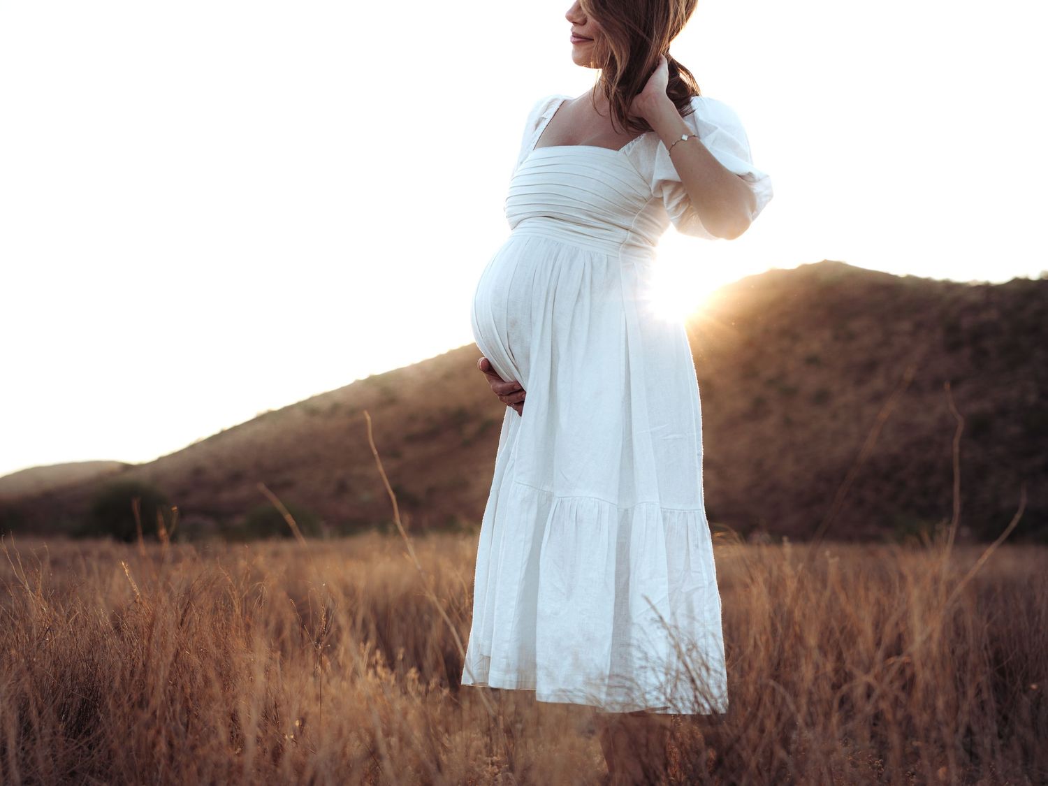 A white maternity dress silhouetted against a sunset in a field of tall grass.