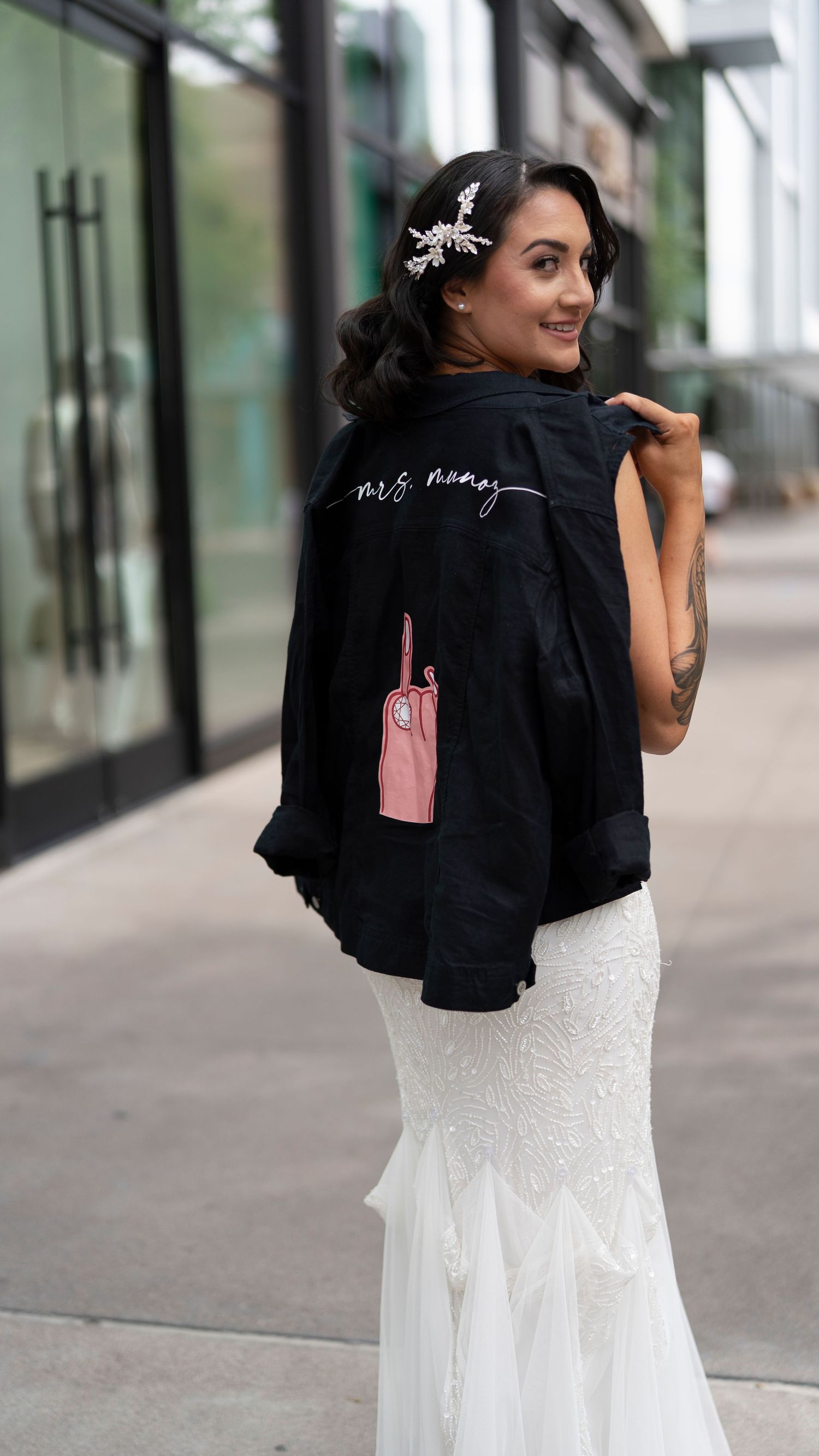 Person wearing a black jacket with white text and a floral hair clip poses in a bridal gown.