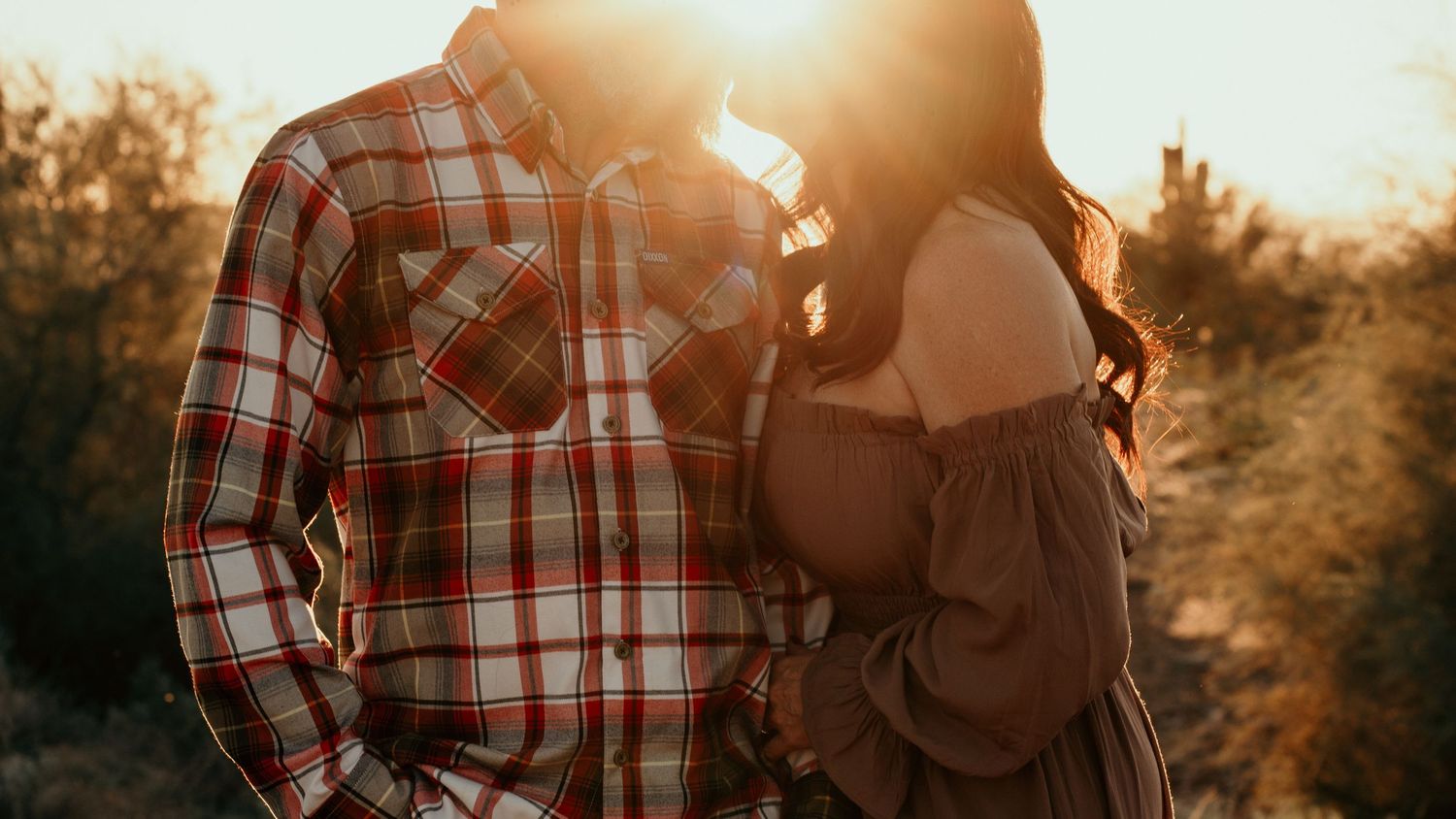 A couple in plaid shirts embrace in warm sunset light surrounded by autumn trees and golden sunflare.