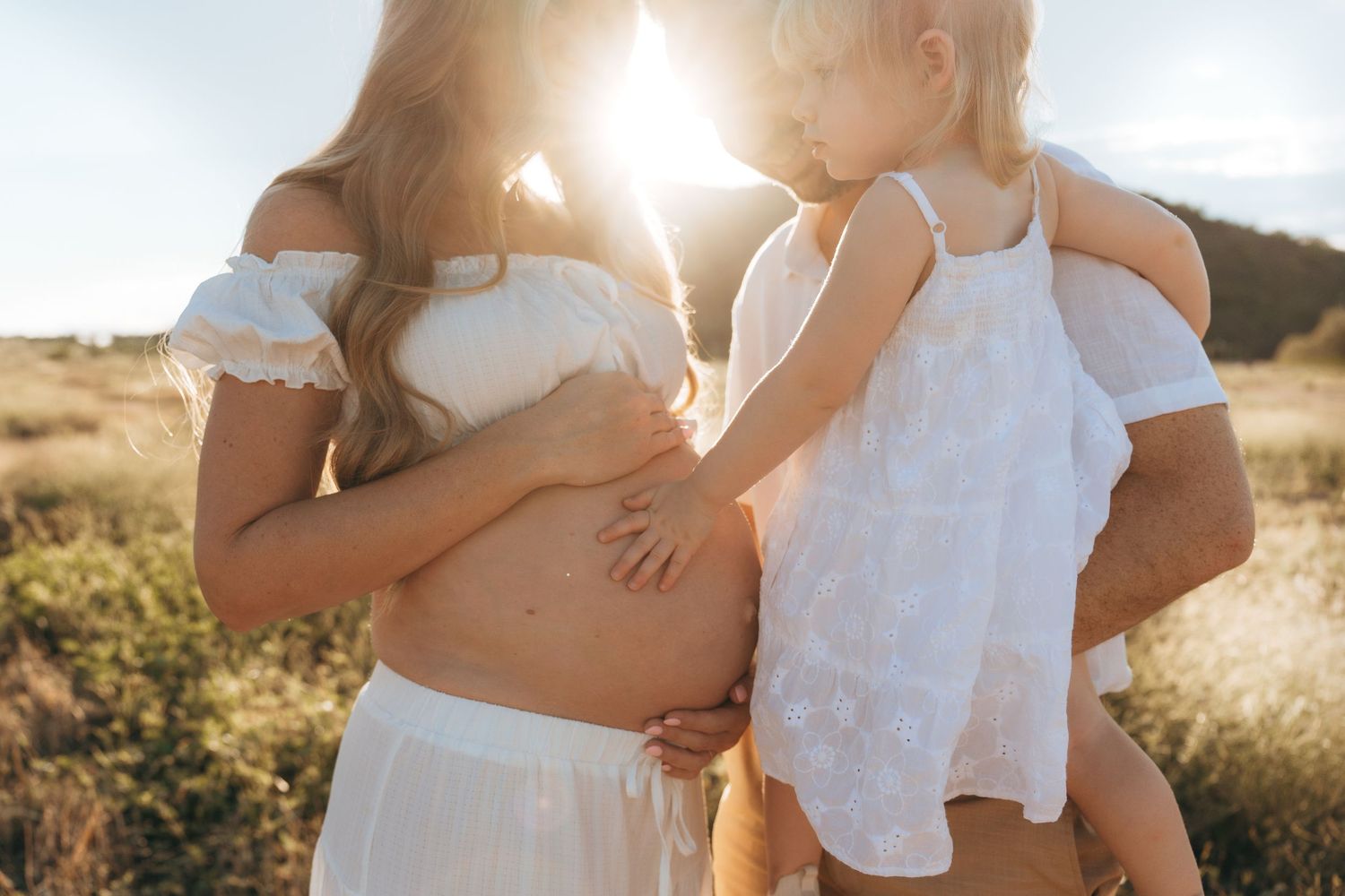 A couple embraces in a sunlit field wearing white outfits during a romantic sunset photoshoot.