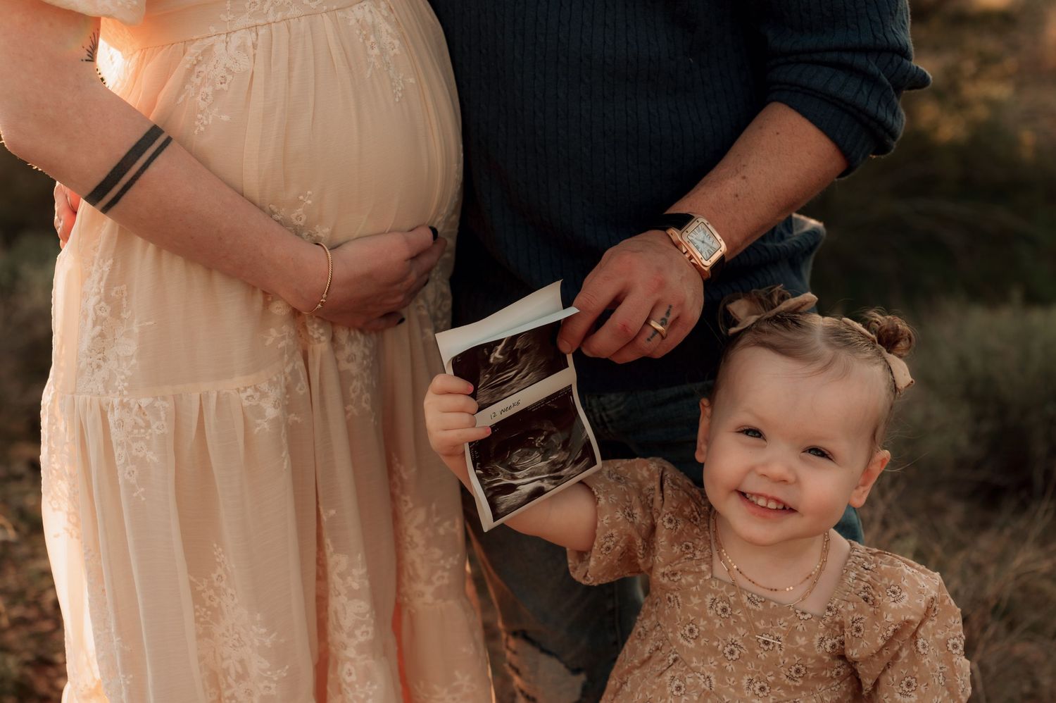 A young child in a pink dress smiles while being held during an outdoor family photoshoot at sunset.