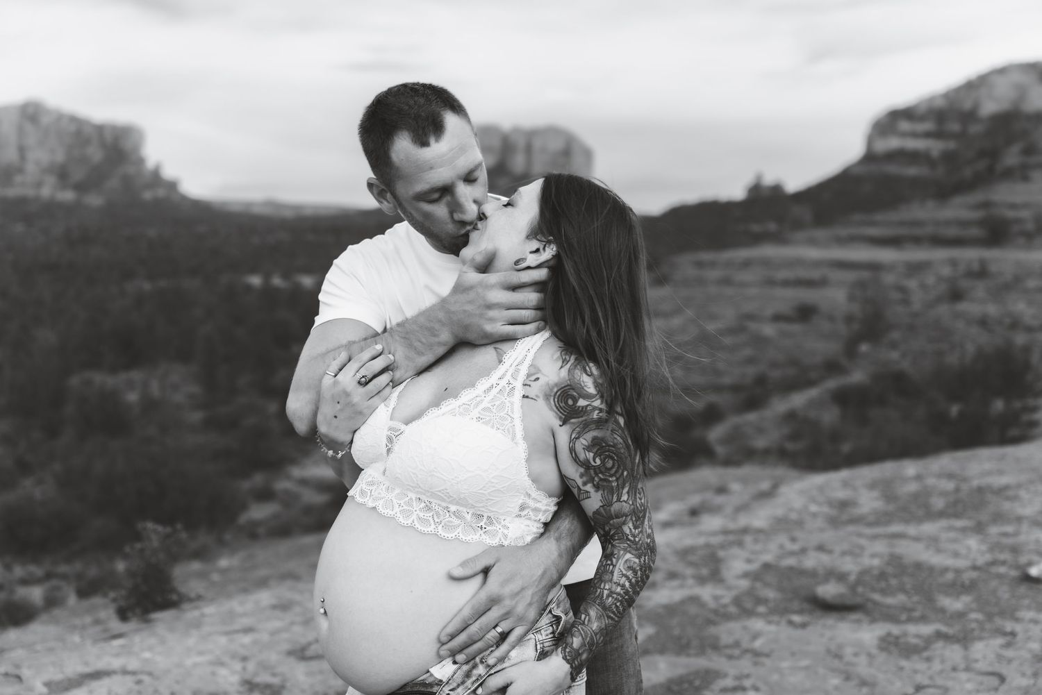 Black and white maternity photo showing couple embracing on rocky terrain with desert landscape in background.