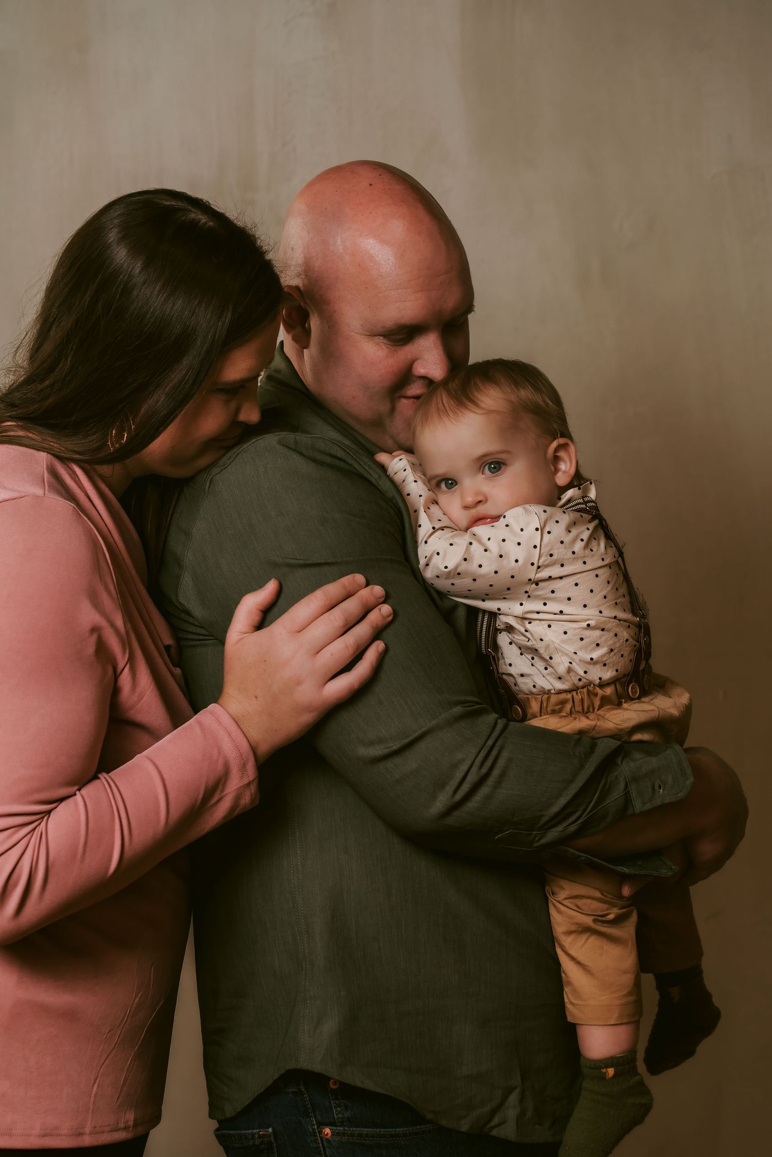 A bald man in a green shirt holds a baby while a woman in pink embraces them in a warm family portrait.
