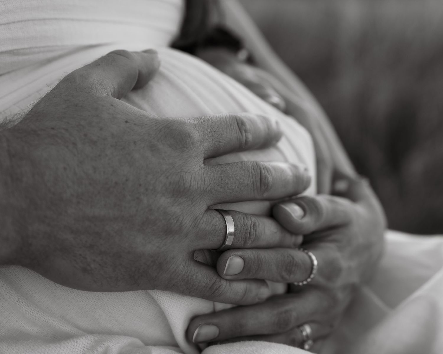 Close-up black and white photo of hands with wedding rings resting on maternity dress.