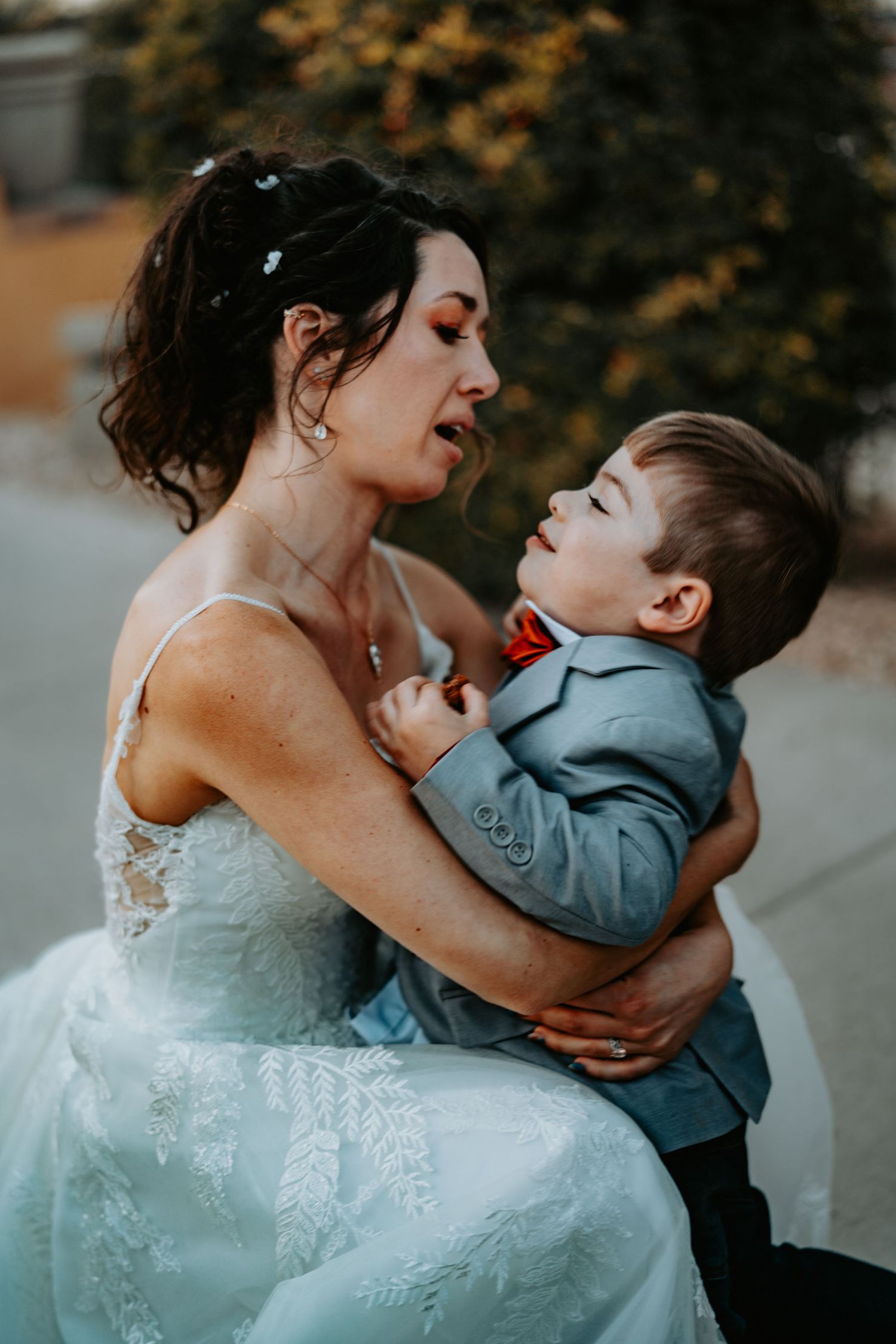 Person in white wedding gown embraces small child in gray suit during emotional moment.