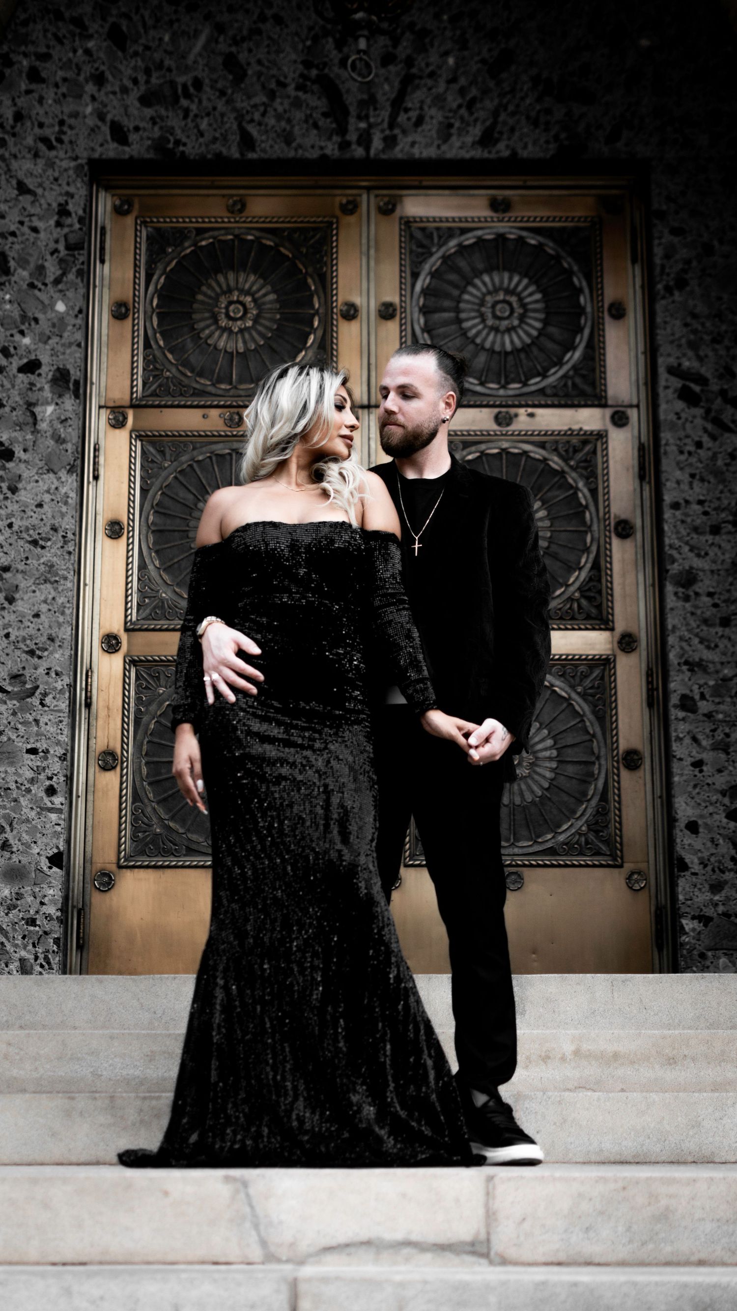 A romantic couple poses in formal attire against an ornate backdrop with decorative doors.