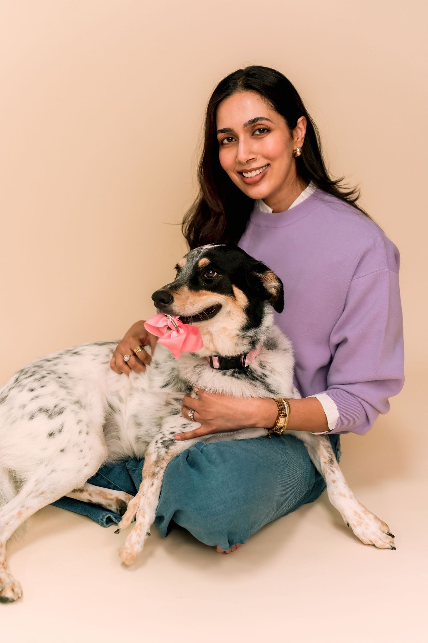 A person in a purple sweater sits with their spotted black and white rescue dog wearing a pink collar against a beige backdrop.